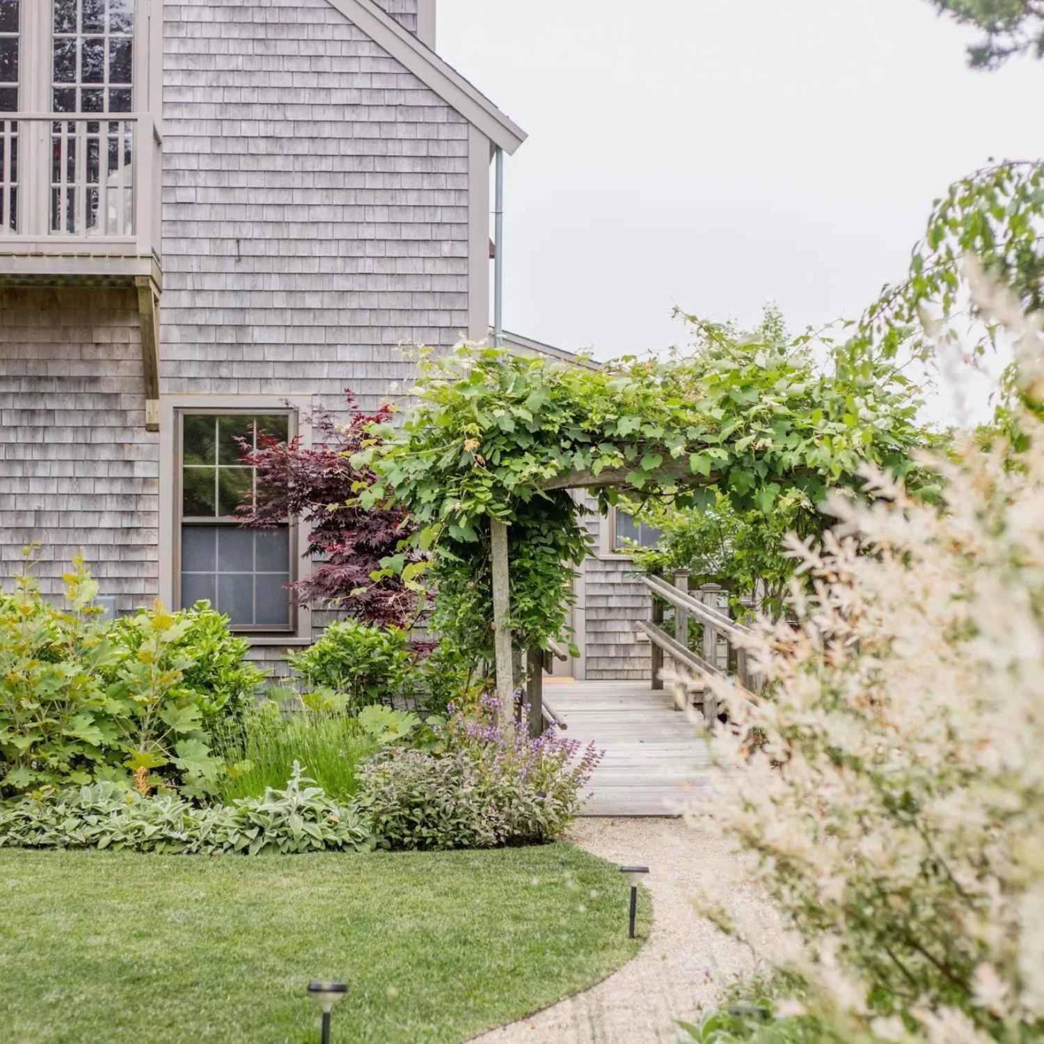 Exterior of Nantucket Lighthouse School’s Middle School campus at 49 Fairgrounds Road, featuring a traditional gray-shingled building surrounded by lush greenery and a wooden walkway leading to the entrance beneath a vine-covered trellis.