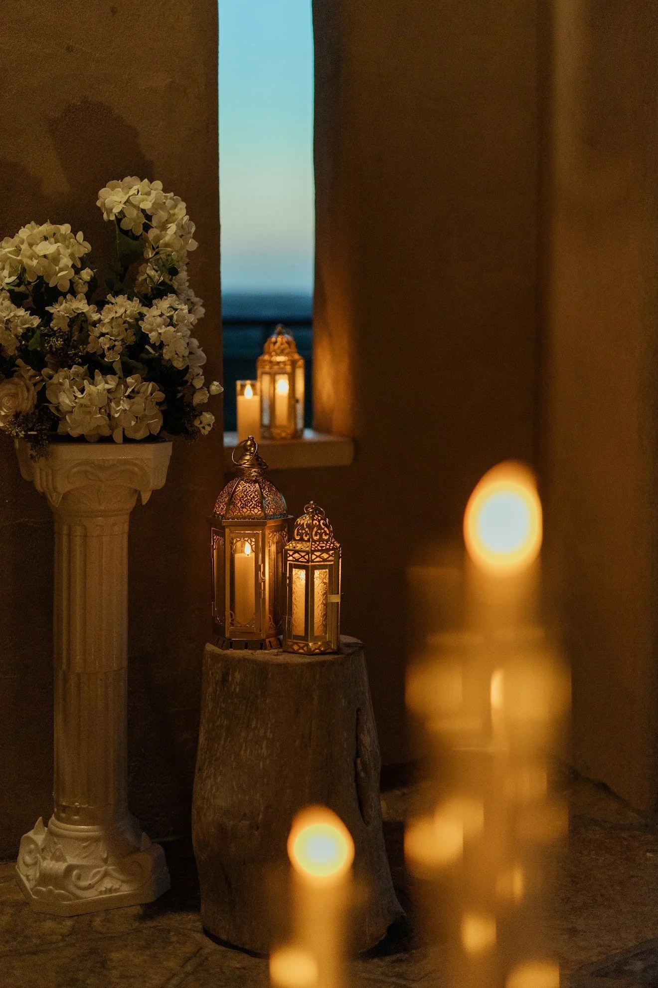 A tall white floral arrangement on a pedestal, lit by two Moroccan-style lanterns and a candle on a wooden post, with a view of the ocean and a lantern on a windowsill in the background at dusk.