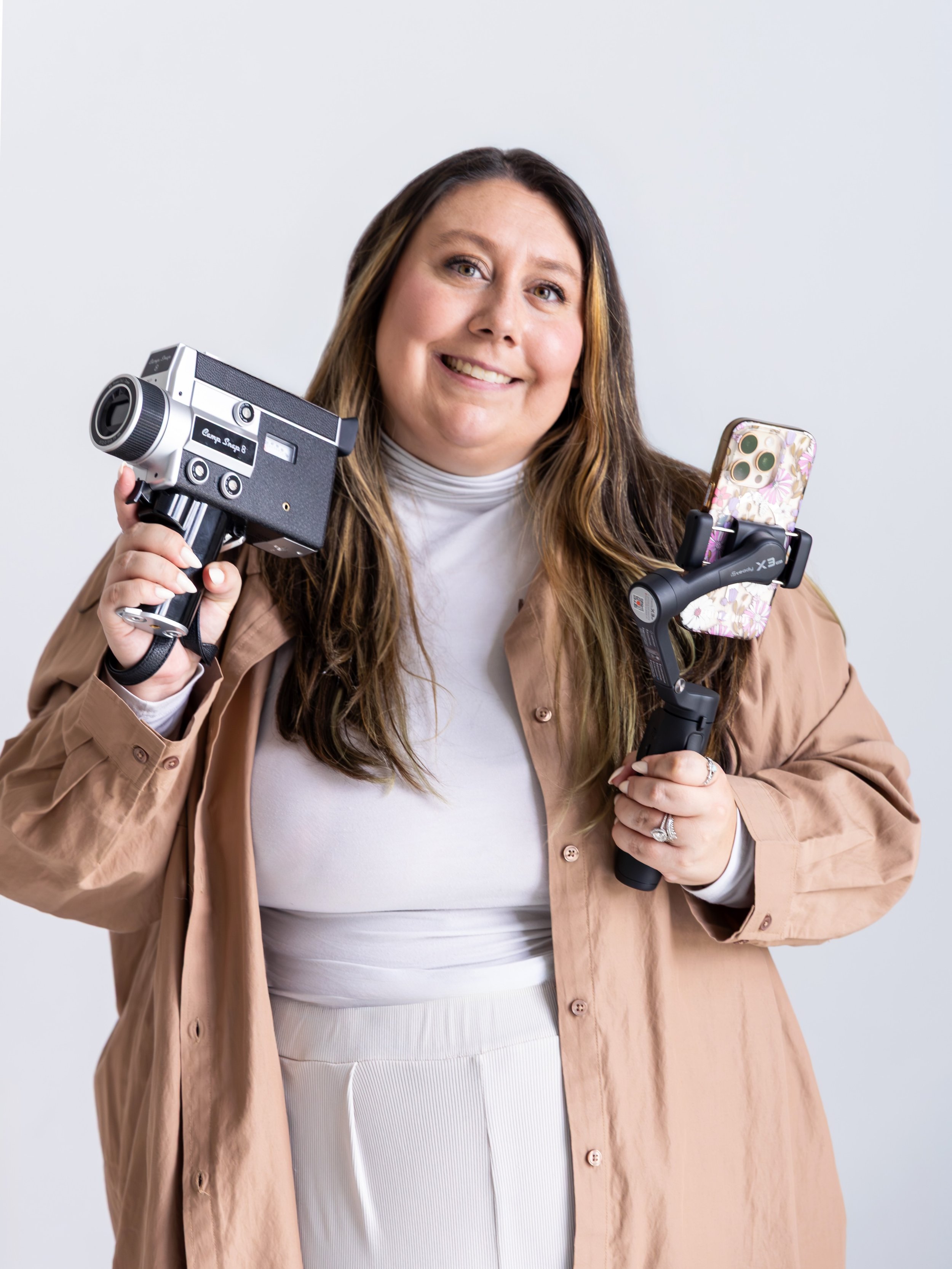 A woman with long brown hair smiling while holding a vintage camera in her right hand and a smartphone mounted on a stabilizer in her left hand, against a plain light background.