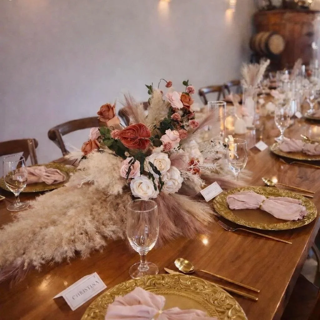 A decorated dining table with a floral centerpiece, gold-rimmed plates with pink napkins, gold flatware, and empty wine glasses.