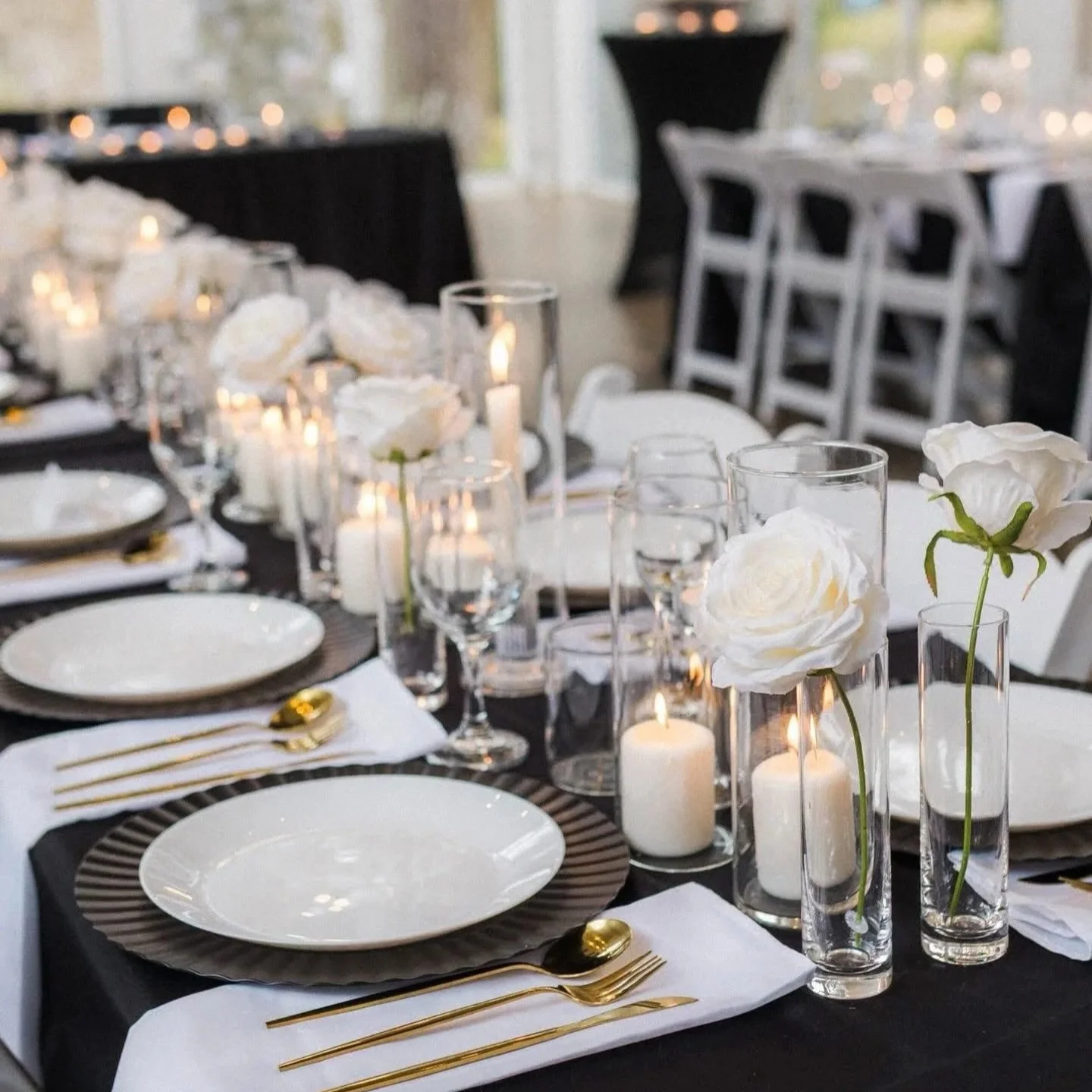 Elegant dining table setup with white roses in tall glass vases, candles, white plates, gold flatware, and black-and-white napkins, ready for a formal event.