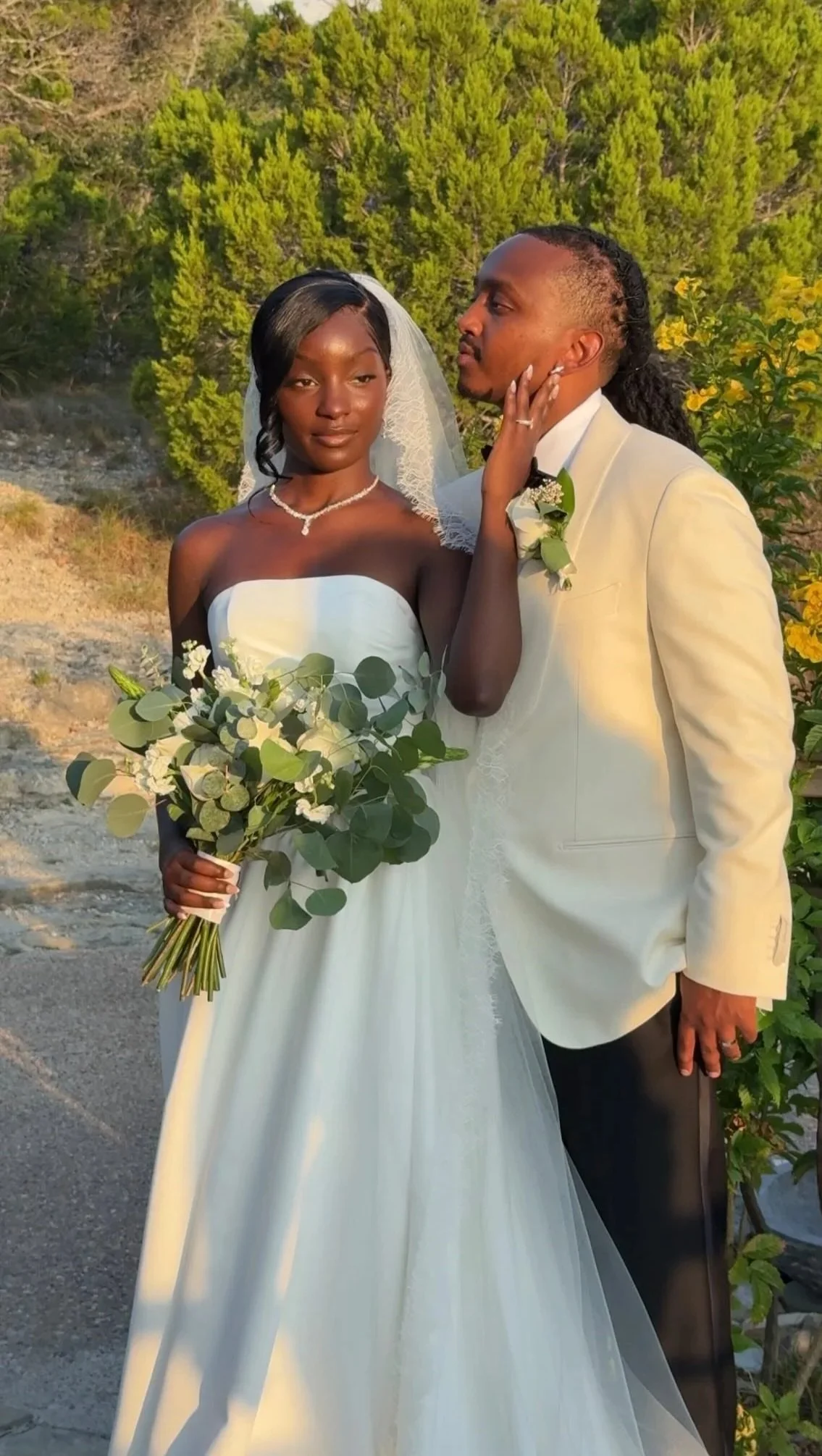 A bride and groom standing outdoors with green trees and bushes in the background during sunset. The bride is holding a bouquet of greenery and white flowers, wearing a strapless white wedding gown, veil, and pearl jewelry. The groom is dressed in a 
