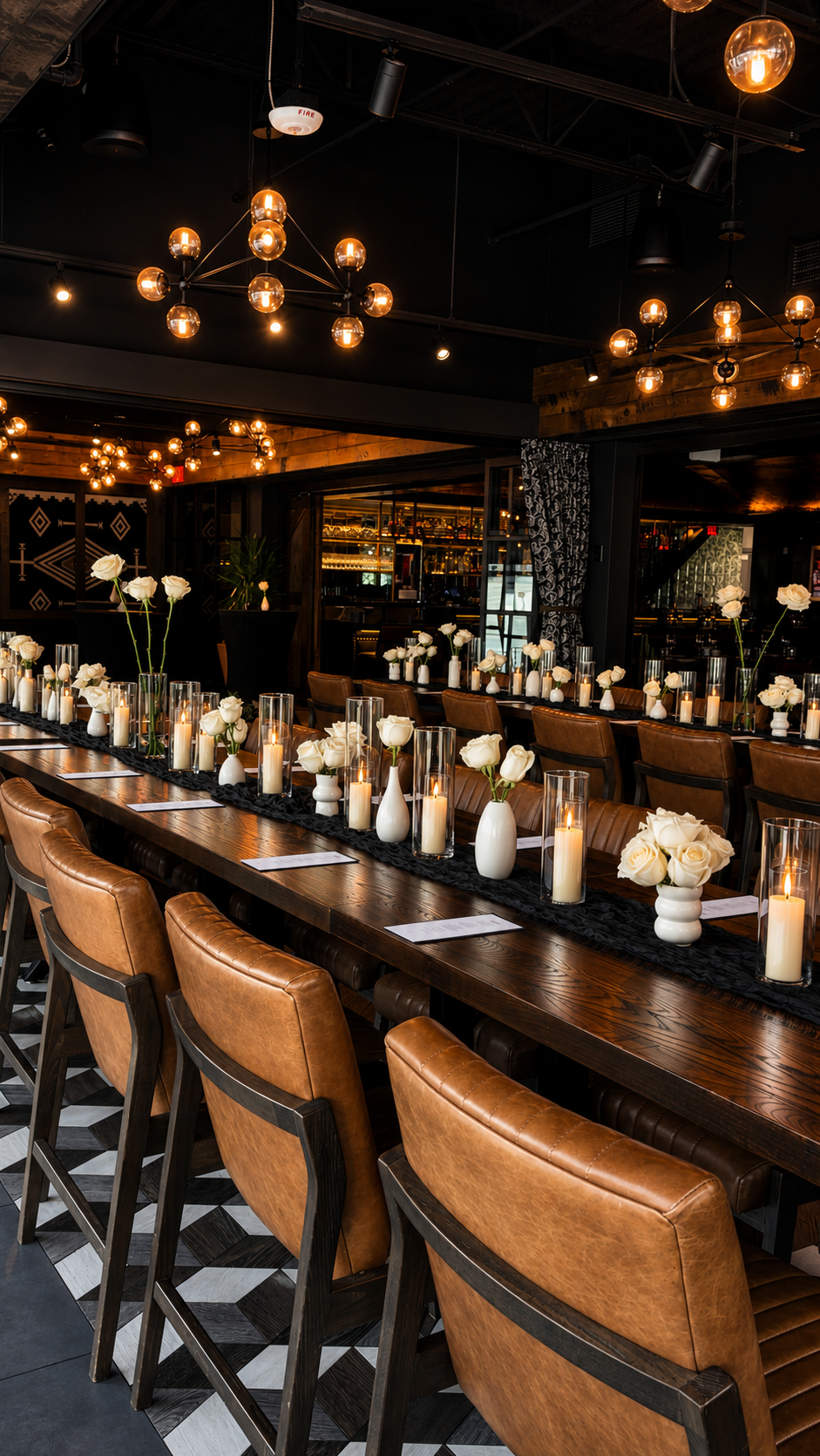An elegantly decorated dining area with a long wooden table, white roses in vases, and tall glass candles. Warm lighting from modern chandeliers illuminates the space, which features a black and brown color scheme and contemporary decor.