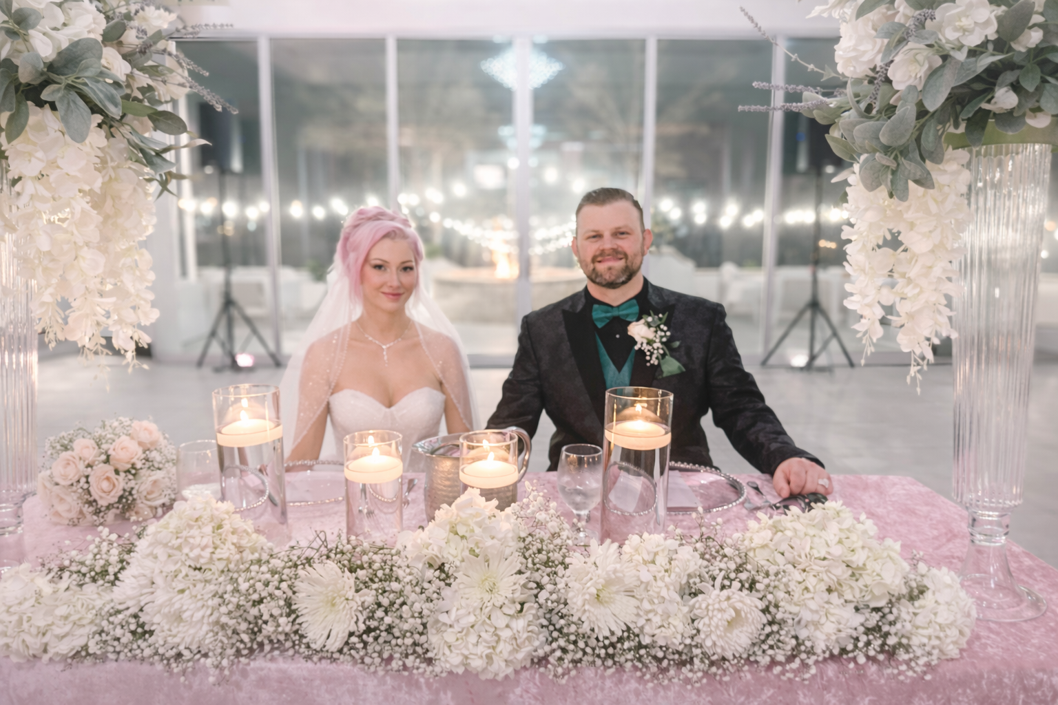 A newlywed couple at their wedding reception seated at a decorated table with white and pink flowers, candles, and elegant tableware. The bride has pink hair and wears a wedding dress, while the groom has a beard and is dressed in a tuxedo with a gre