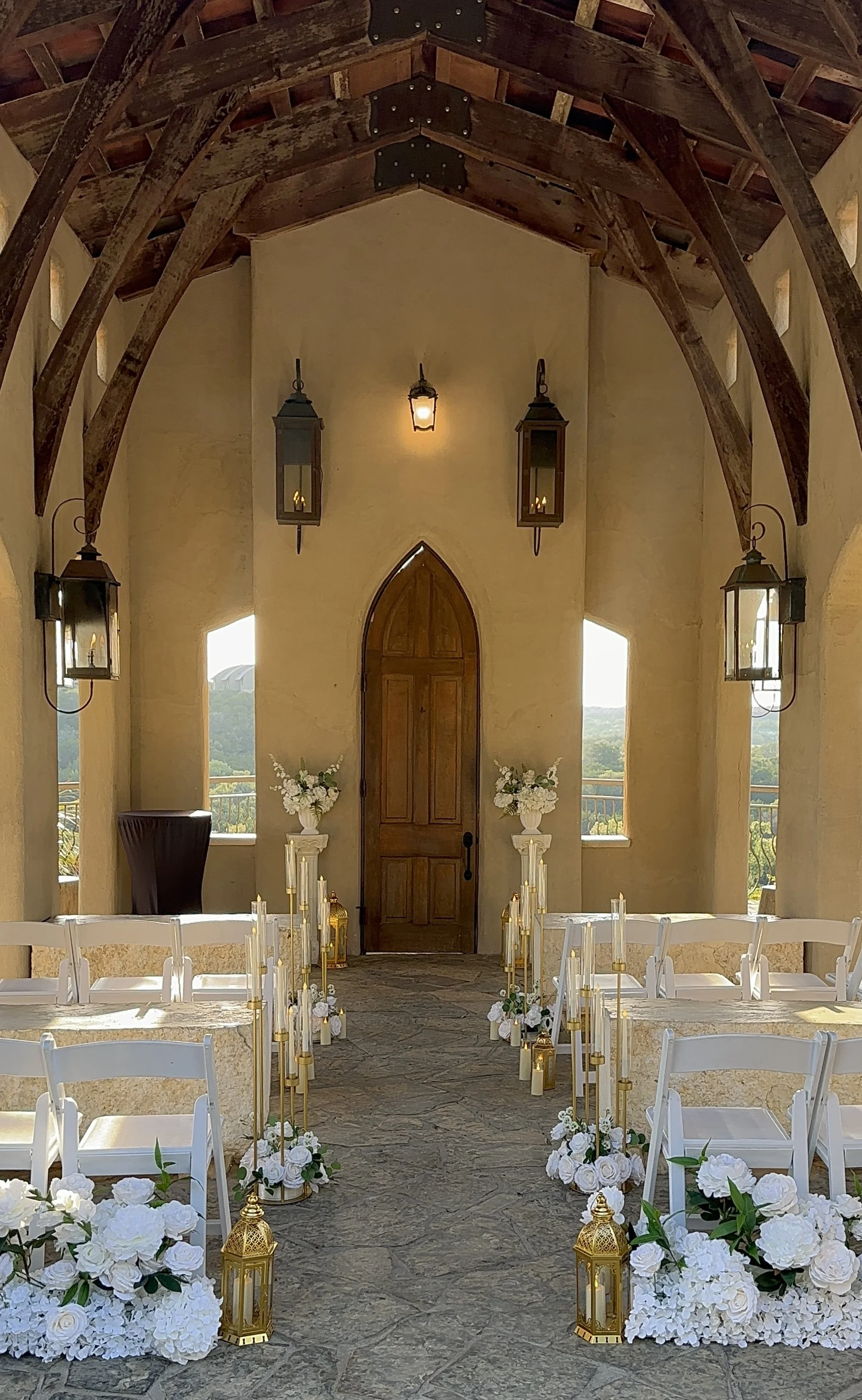 Elegant wedding ceremony setup with white floral arrangements, candles, and gold lanterns, inside a venue with a wooden door, lantern sconces, and a vaulted wooden ceiling.