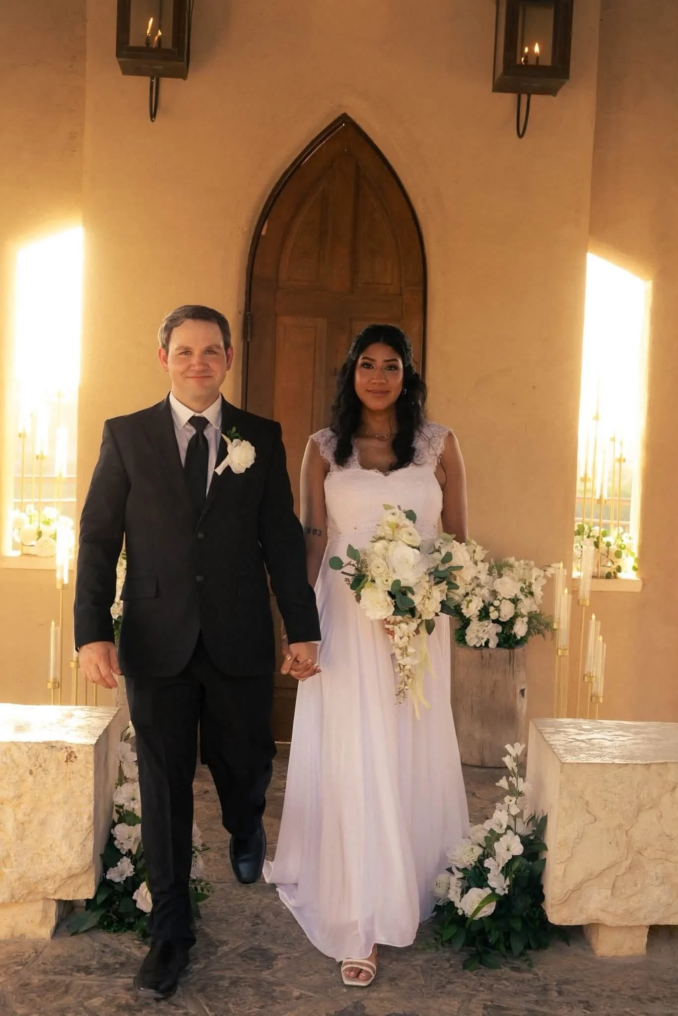 A couple dressed in wedding attire standing hand in hand inside a church, surrounded by white flowers and candles, with warm lighting and an arched wooden door in the background.