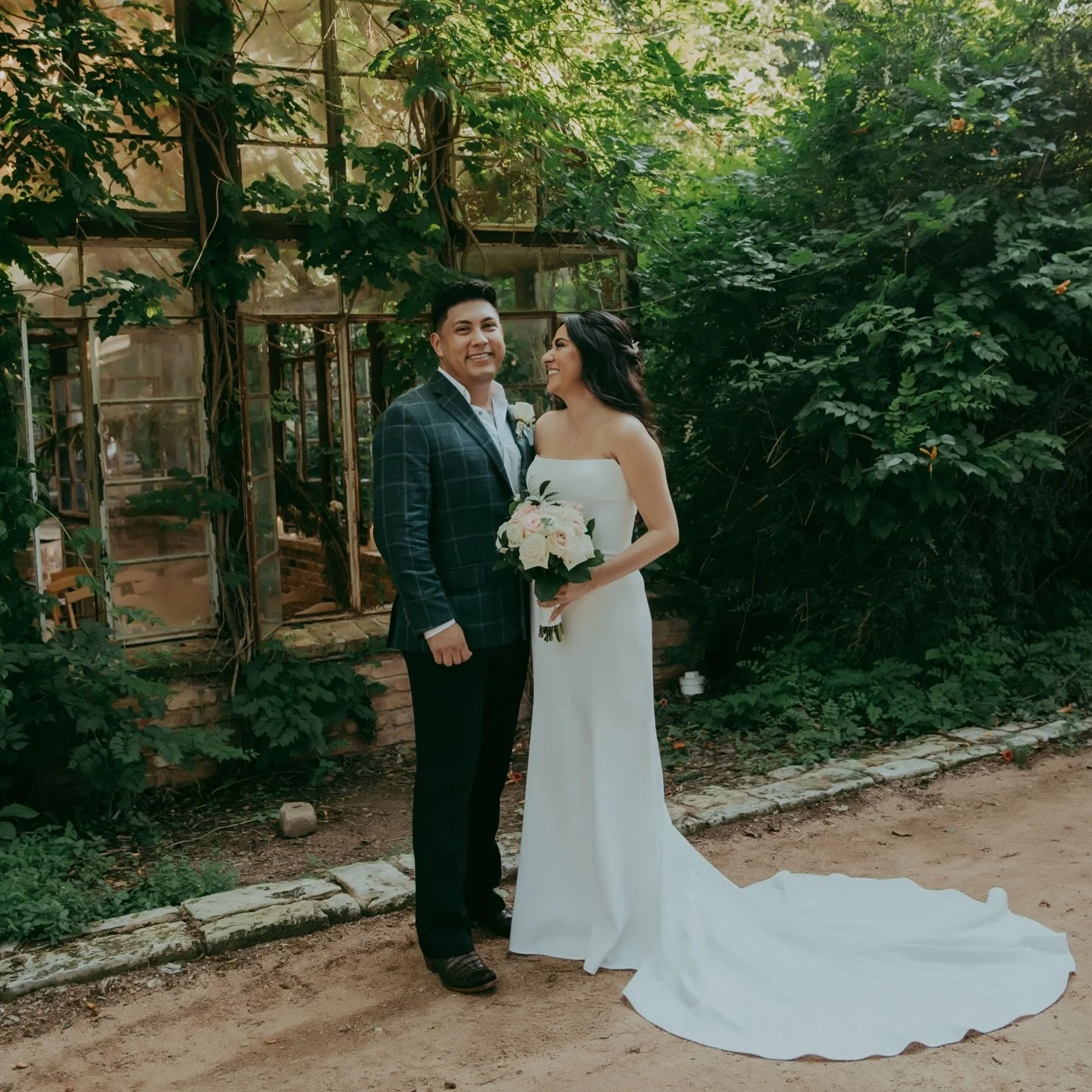 A newlywed couple standing outdoors near a green, leafy background, with the bride holding a bouquet of white and pink roses, both smiling at each other.
