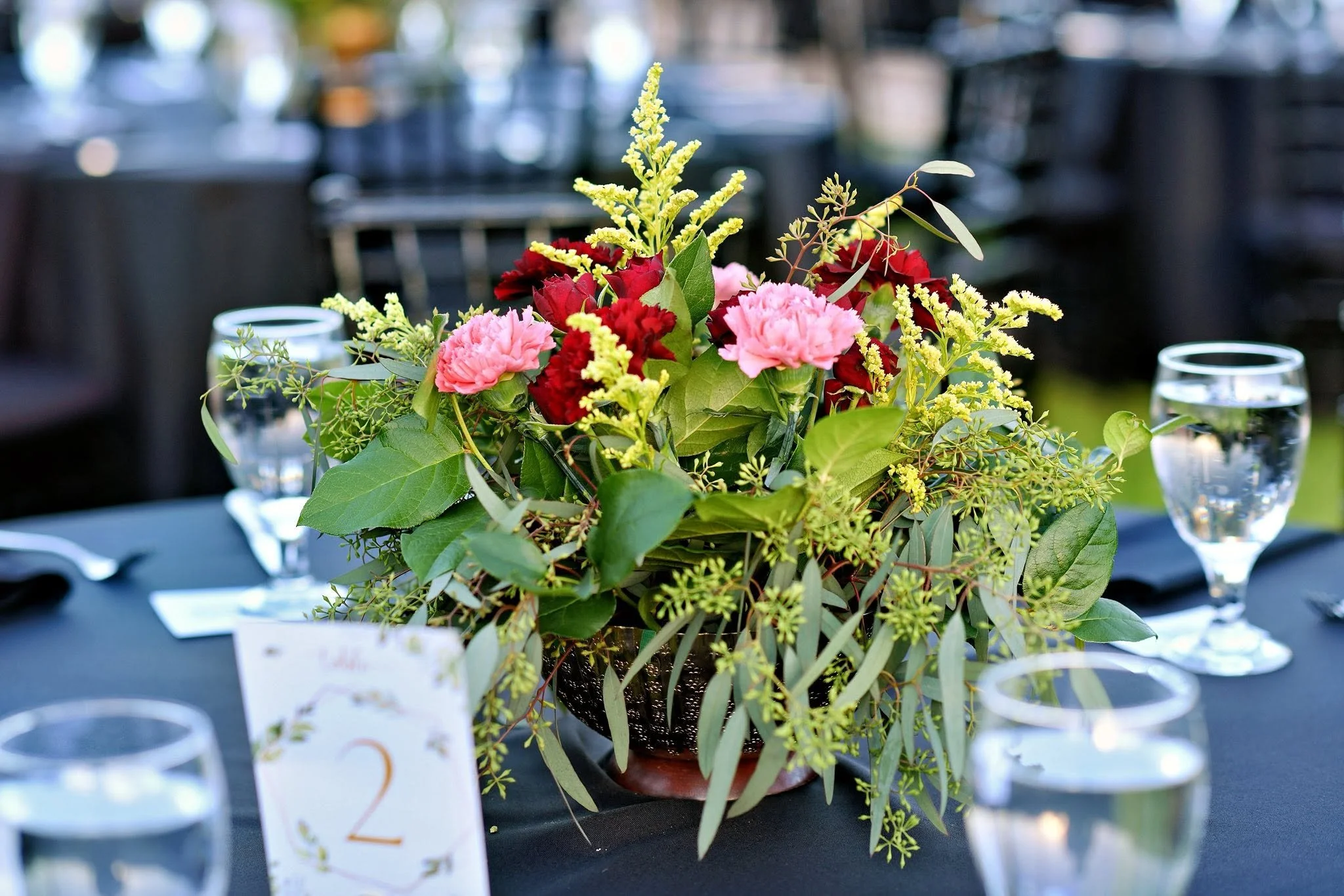 A colorful flower arrangement with pink and red flowers, surrounded by green foliage, placed on a table at an outdoor event with glasses of water in the background.
