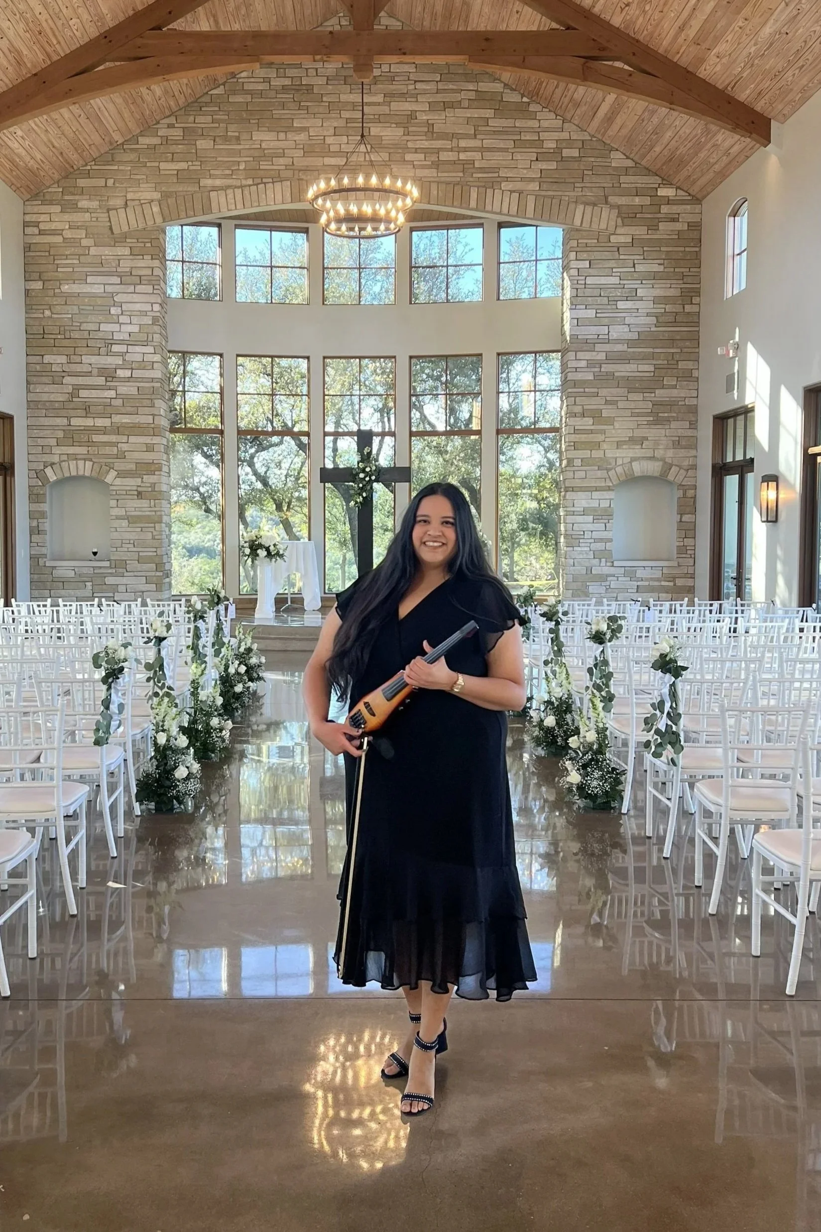 A woman with long dark hair in a black dress and high heels, holding a guitar, posing inside a church decorated for a wedding with a large cross and floral arrangements.