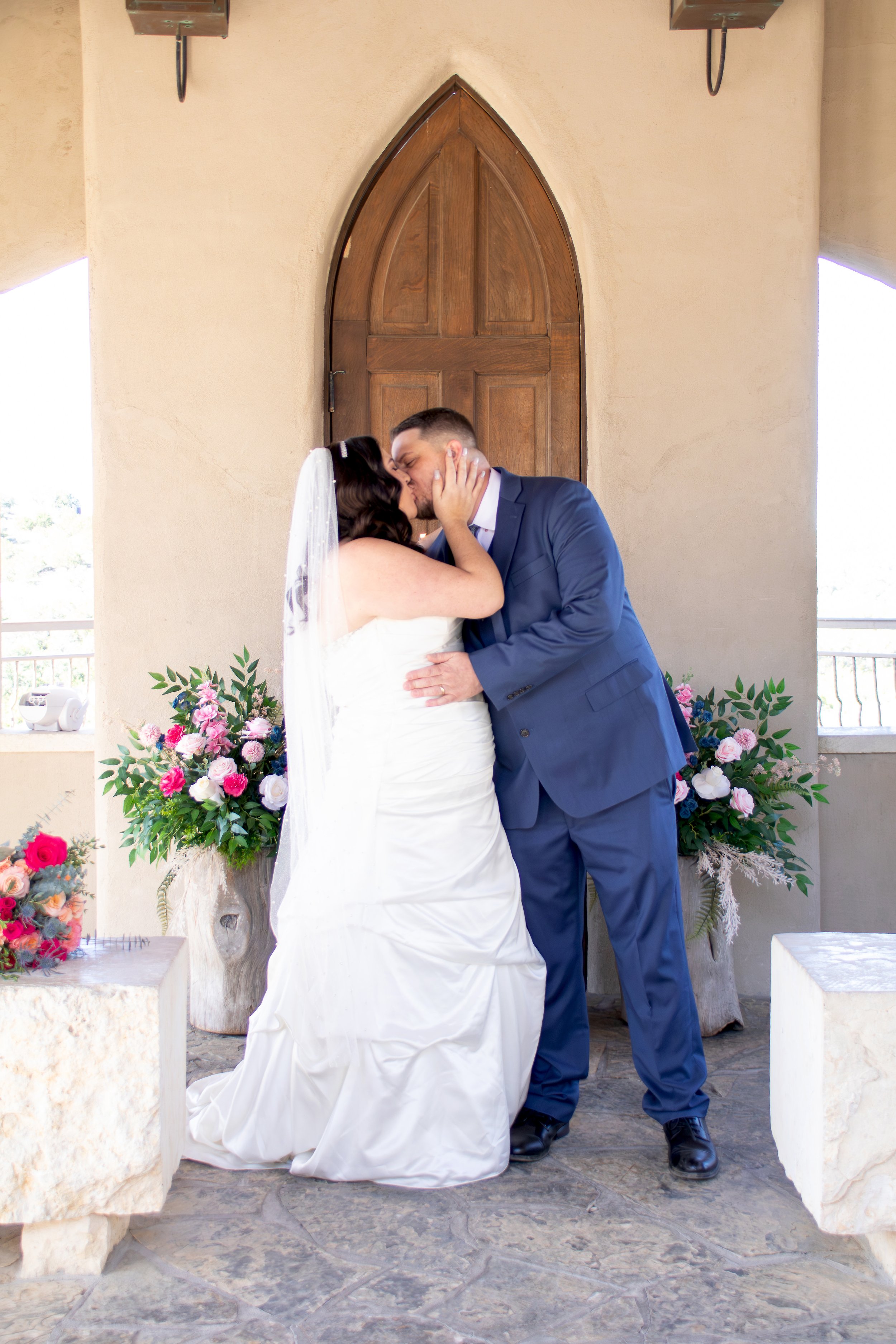 A bride and groom sharing a kiss during their wedding ceremony, standing in front of a wooden arched door with floral arrangements on either side.