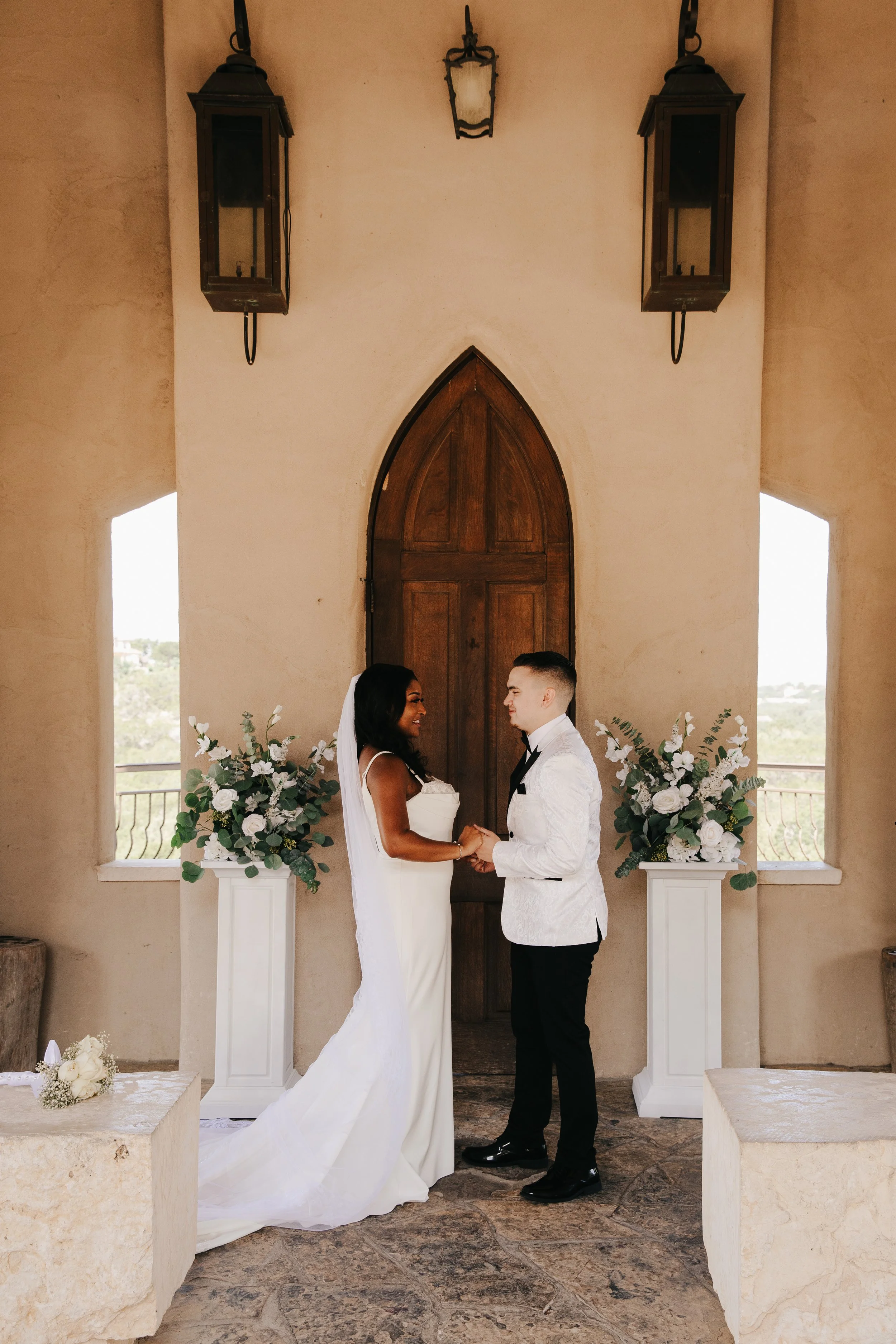 A bride and groom holding hands and facing each other, standing in front of a wooden archway with floral arrangements, inside a building with beige walls and large windows.
