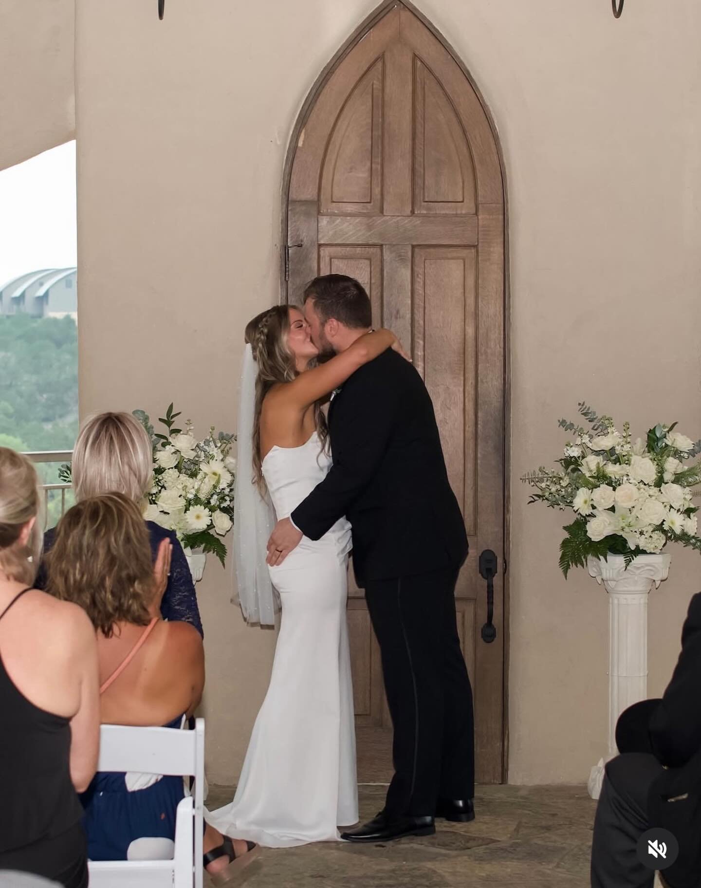 A bride and groom kiss during their wedding ceremony in front of seated guests, with floral arrangements on tall white pedestals on either side of them.