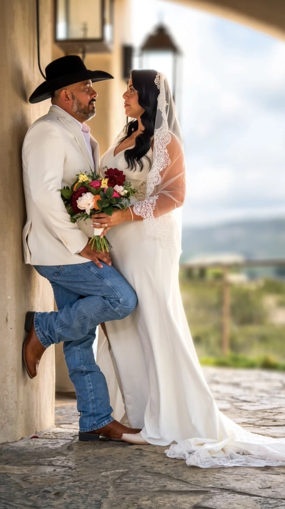 A man in a white blazer, jeans, and cowboy hat and a woman in a white wedding dress and lace veil stand close together outdoors, holding a bouquet of colorful flowers with a scenic view in the background.