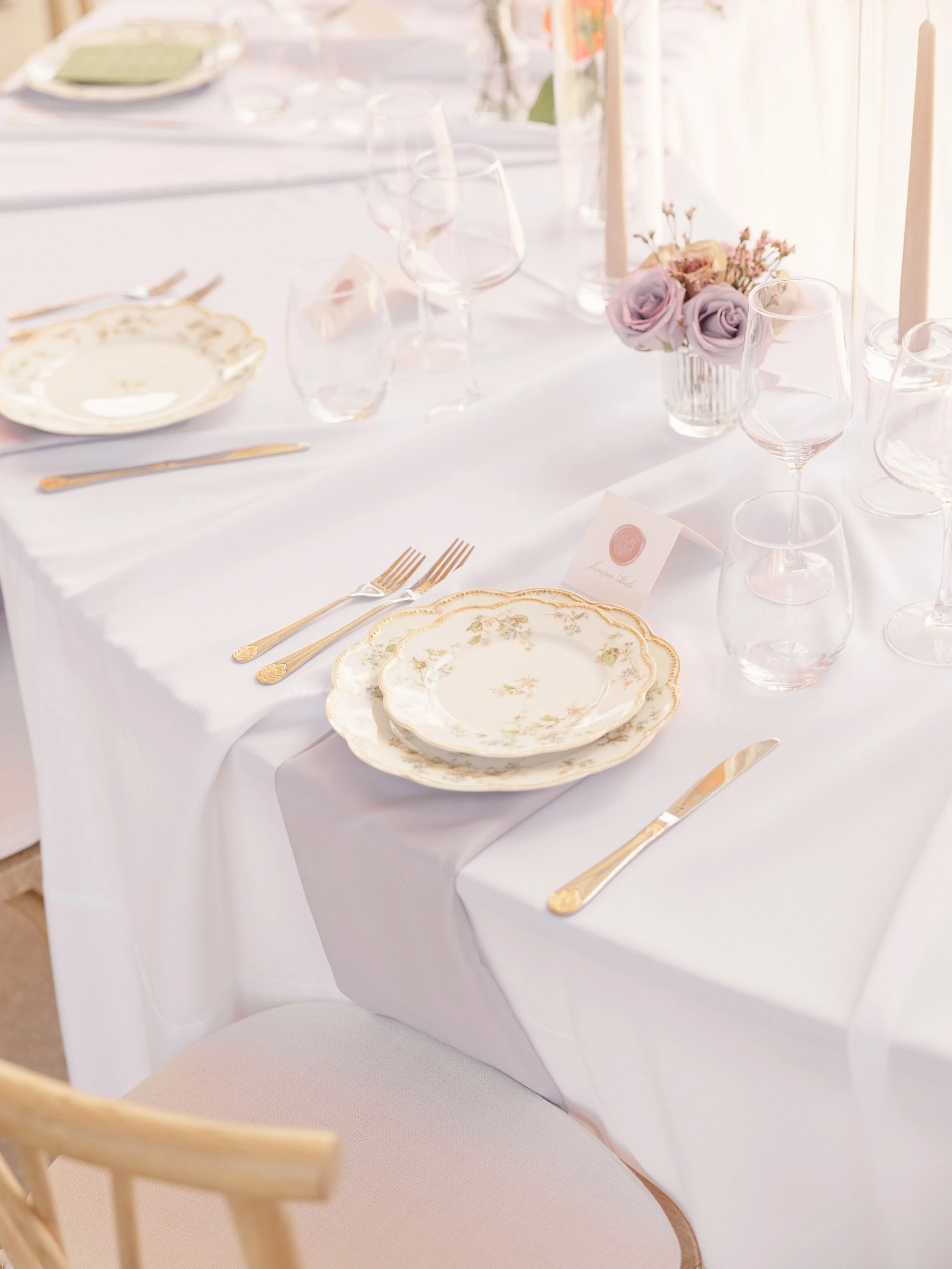 A formal dining table set with vintage floral china plates, gold cutlery, clear glassware, lavender roses in a small vase, tall candles, and a white tablecloth.