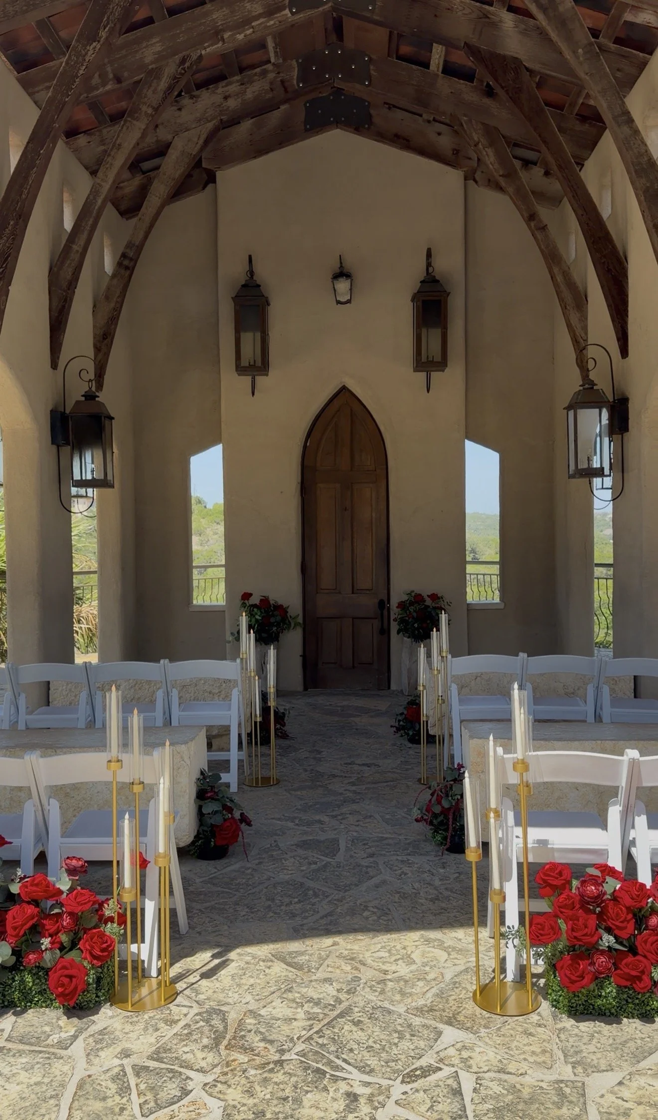 Outdoor wedding ceremony setup with white chairs arranged in rows, gold candleholders with candles, and red roses on the sides. A wooden arched door on a beige wall with three lantern-style lights and view of hills and trees in the background.