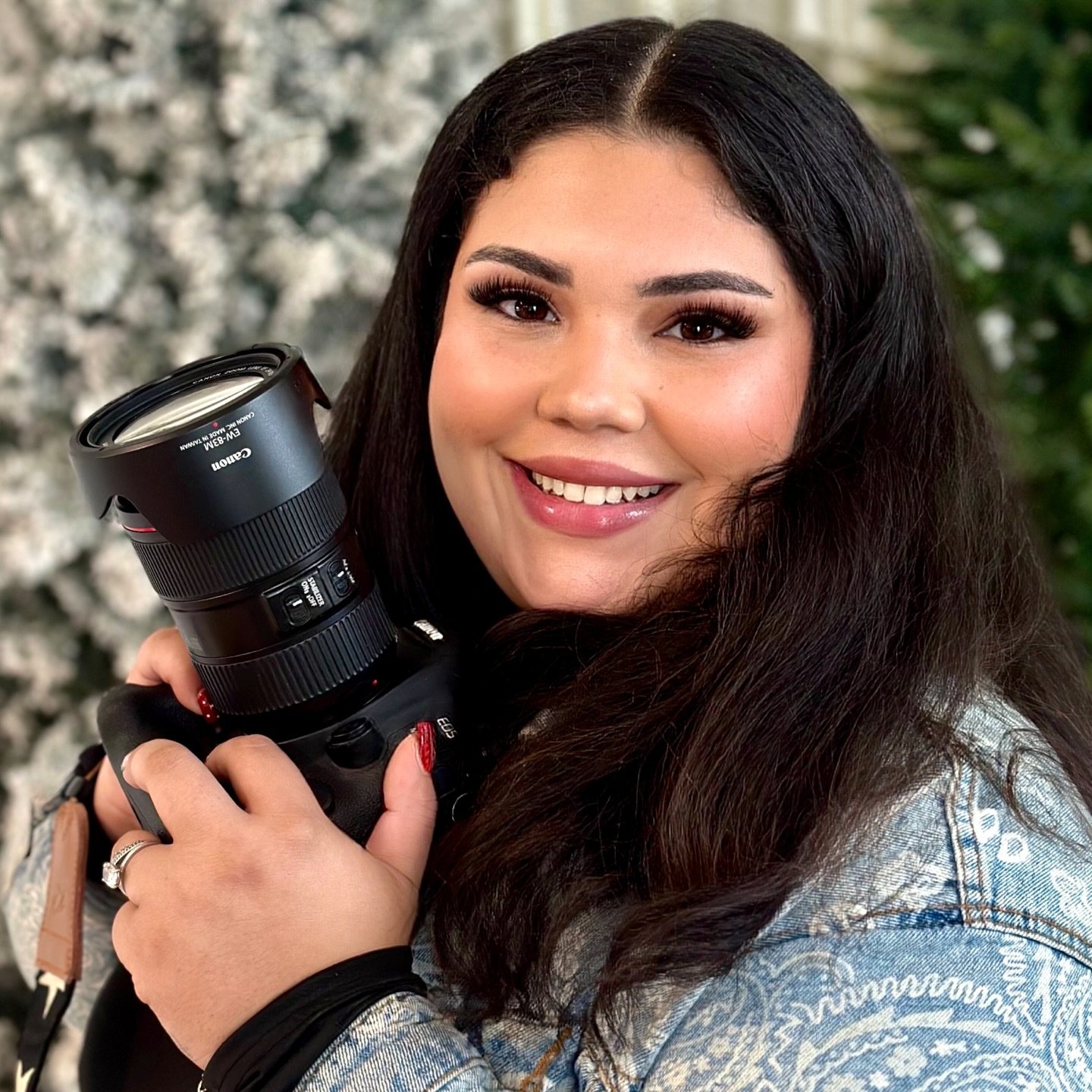 A woman smiling and holding a professional camera outdoors, with a blurred background of trees and flowers.