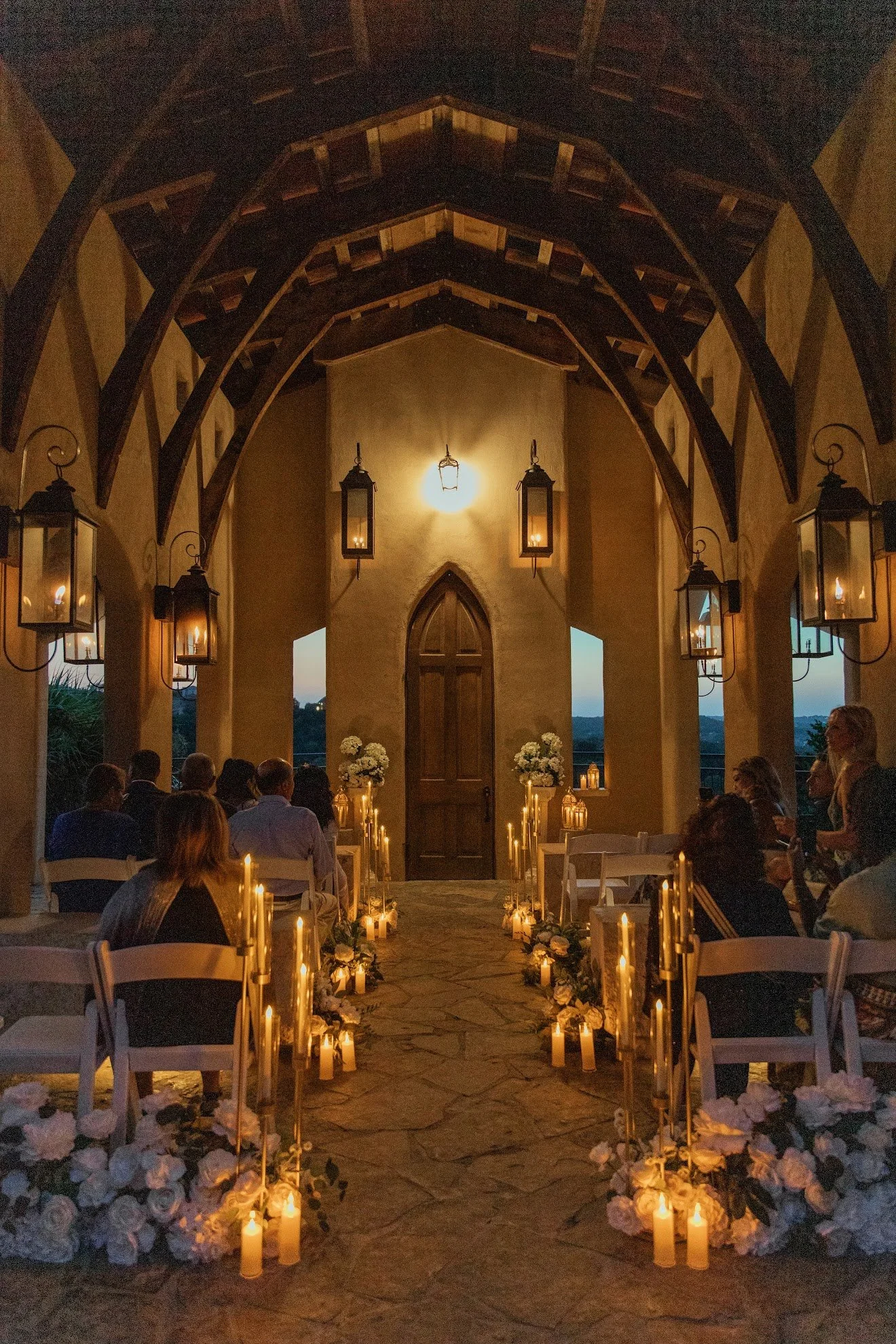 Indoor wedding ceremony with guests seated, candles, and floral arrangements. A wooden door and arched ceiling with hanging lanterns are visible, with dusk or evening light outside.