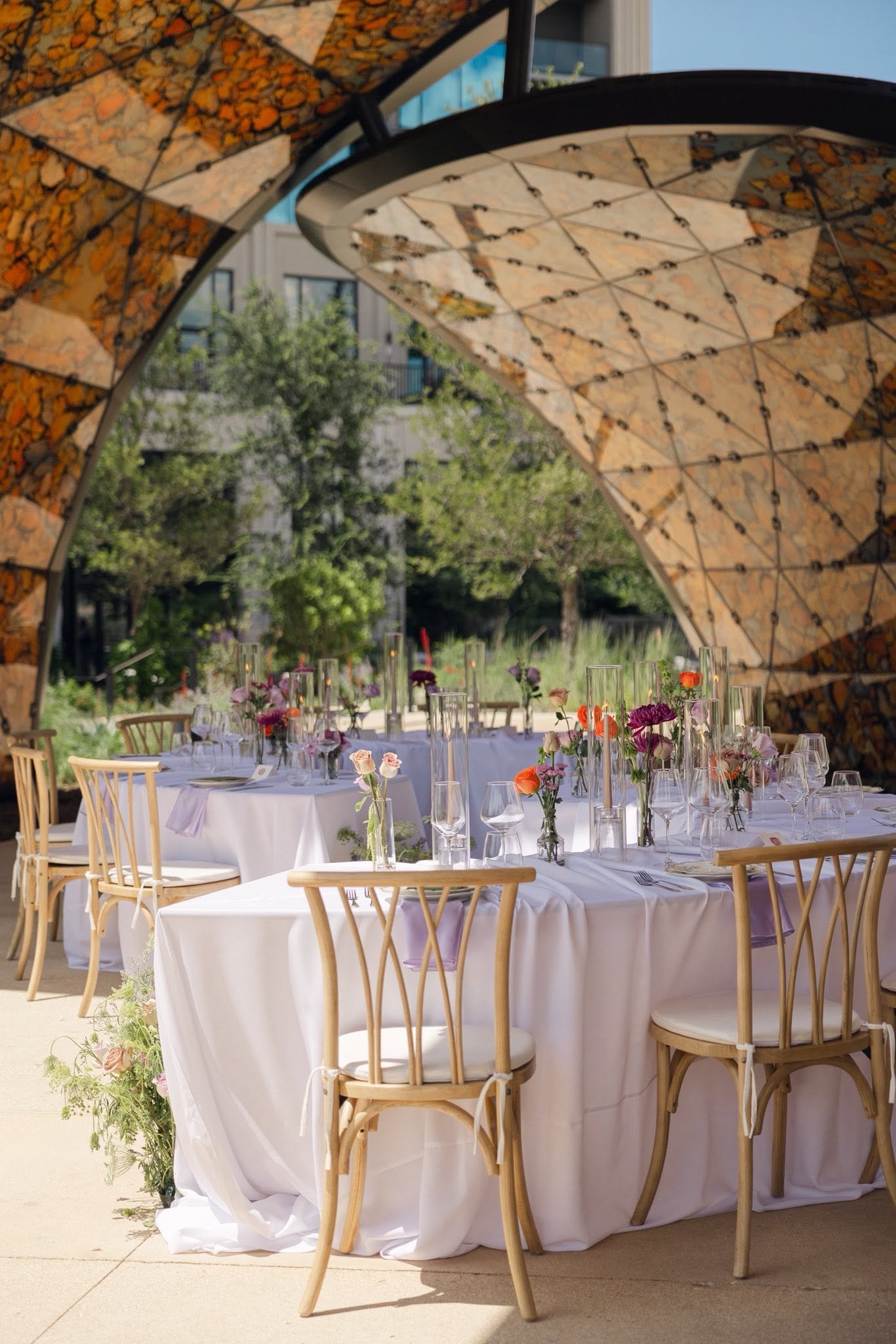 Outdoor dining setup with round tables draped in white cloths, adorned with colorful flower centerpieces in glass vases, surrounded by wooden chairs. The setting is beneath an architectural canopy made of mosaic tiles with earthy tones, with a backgr