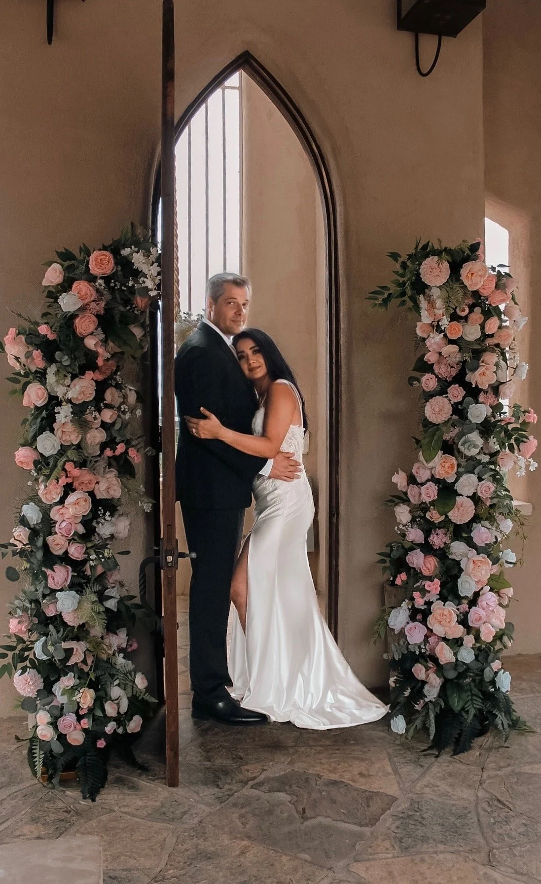 A bride and groom standing in front of a church doorway decorated with pink and white flowers, embracing each other on their wedding day.