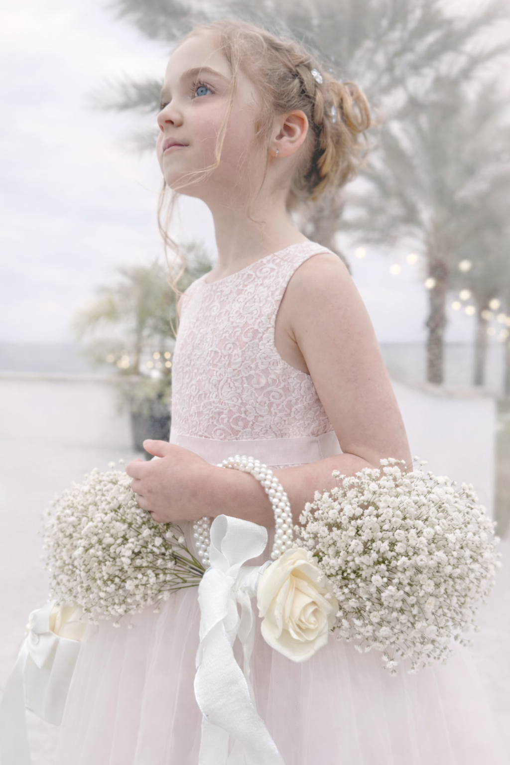 A young girl in a pink lace dress holding a bouquet of white flowers and pearls at an outdoor event.
