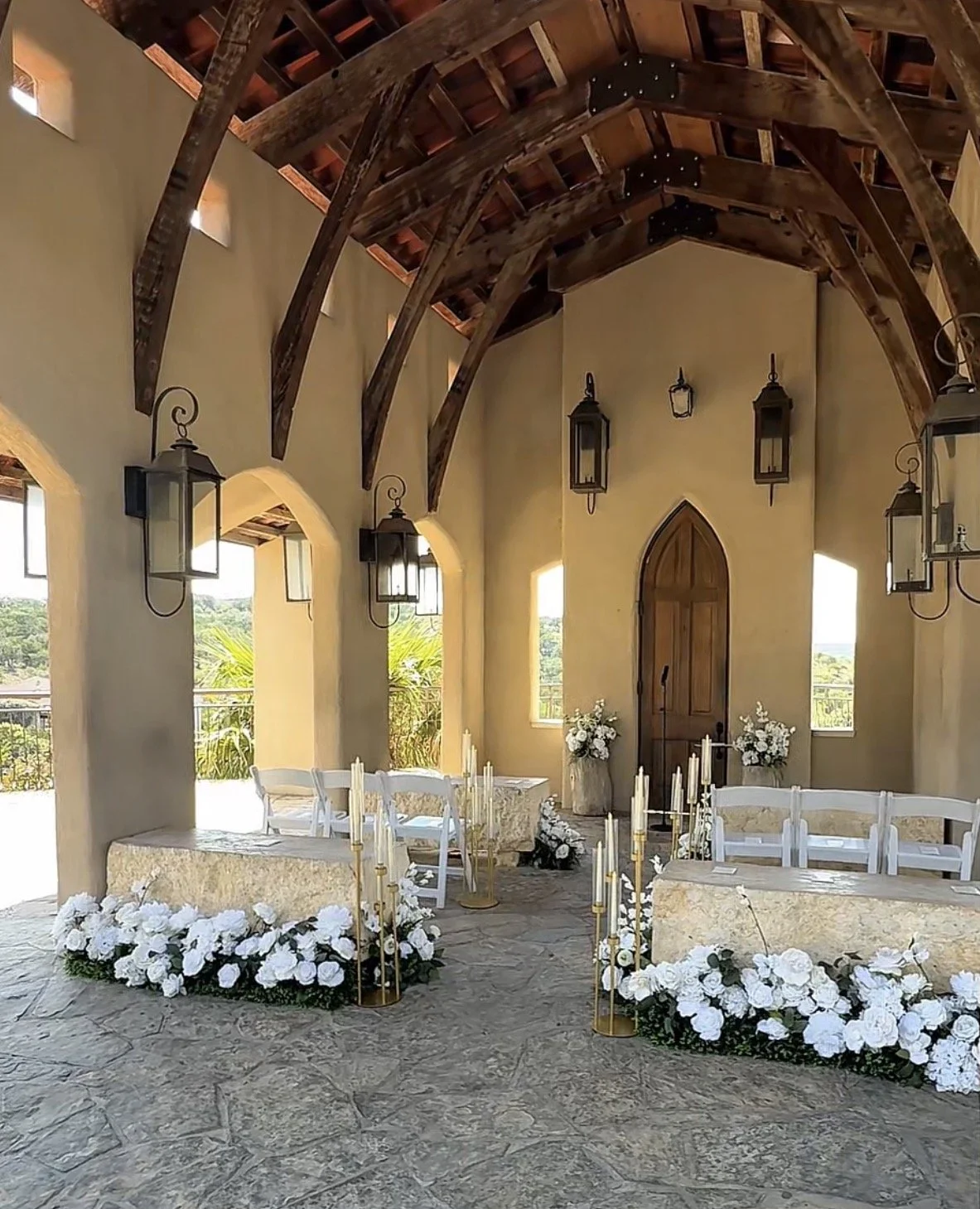 Interior of a chapel decorated for a wedding with white flowers, candles, and white chairs, featuring wooden beams and lanterns on the walls.