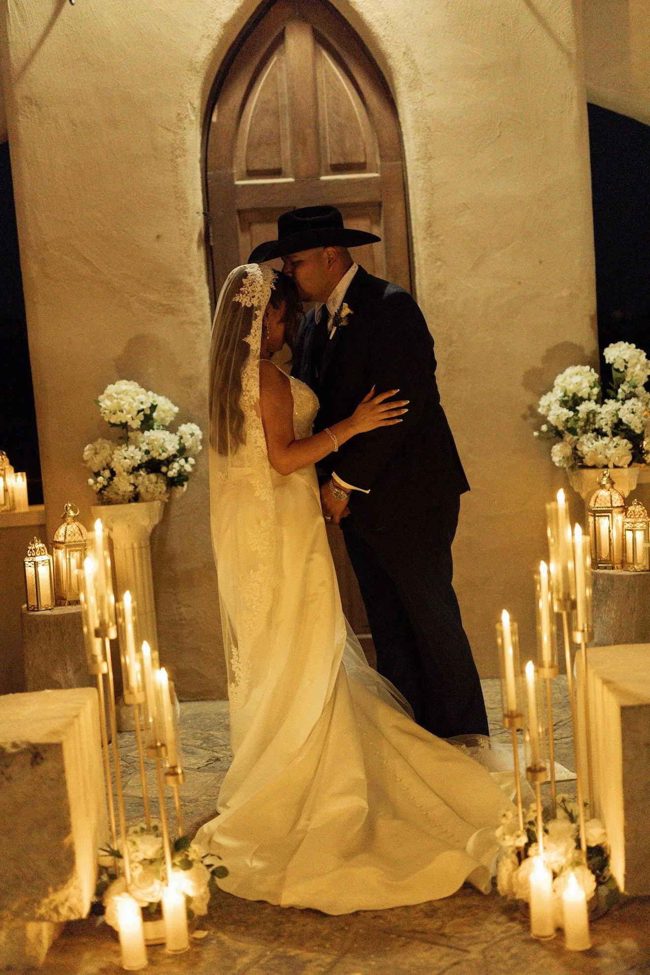 A bride and groom kissing during their wedding ceremony inside a church, surrounded by lit candles and floral arrangements.