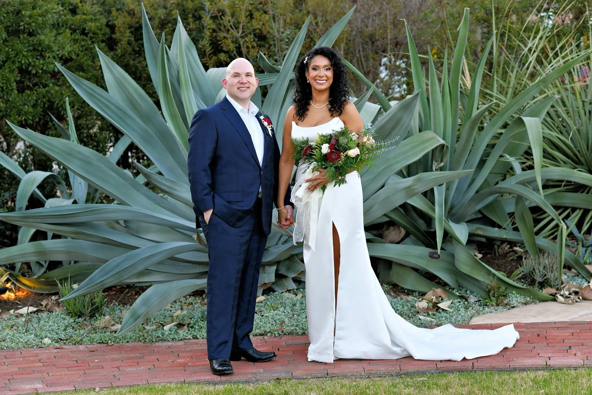 A newlywed couple holding hands outdoors in front of a large agave plant, smiling, dressed in wedding attire with the groom in a navy suit and the bride in a white gown holding a bouquet.