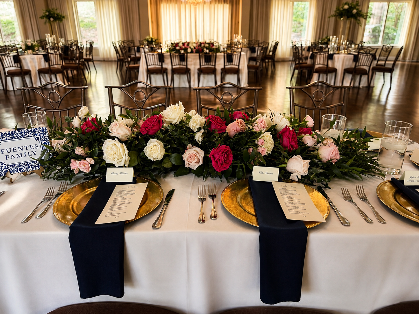 Decorated banquet table with gold plates and black napkins, surrounded by chairs, with a large floral centerpiece of roses and greenery, in an elegant venue with large windows and draped curtains.