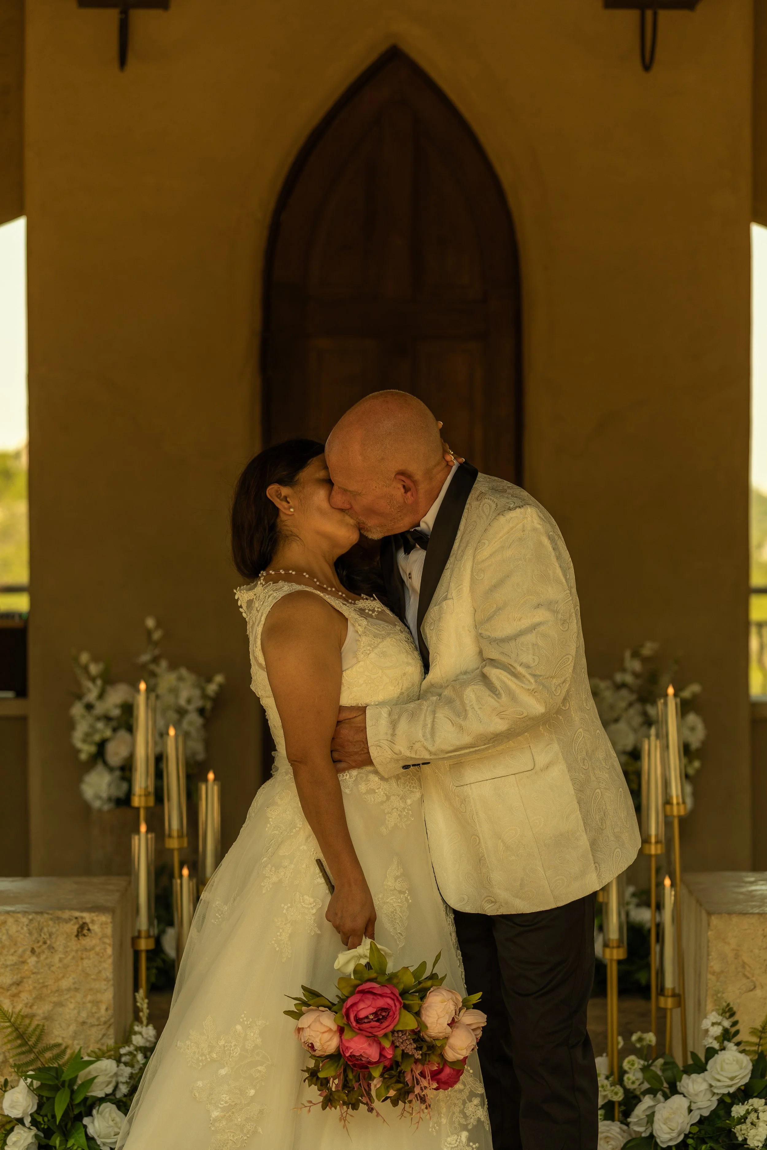 A bride and groom share a kiss during their wedding ceremony inside a chapel with candles and floral arrangements in the background.