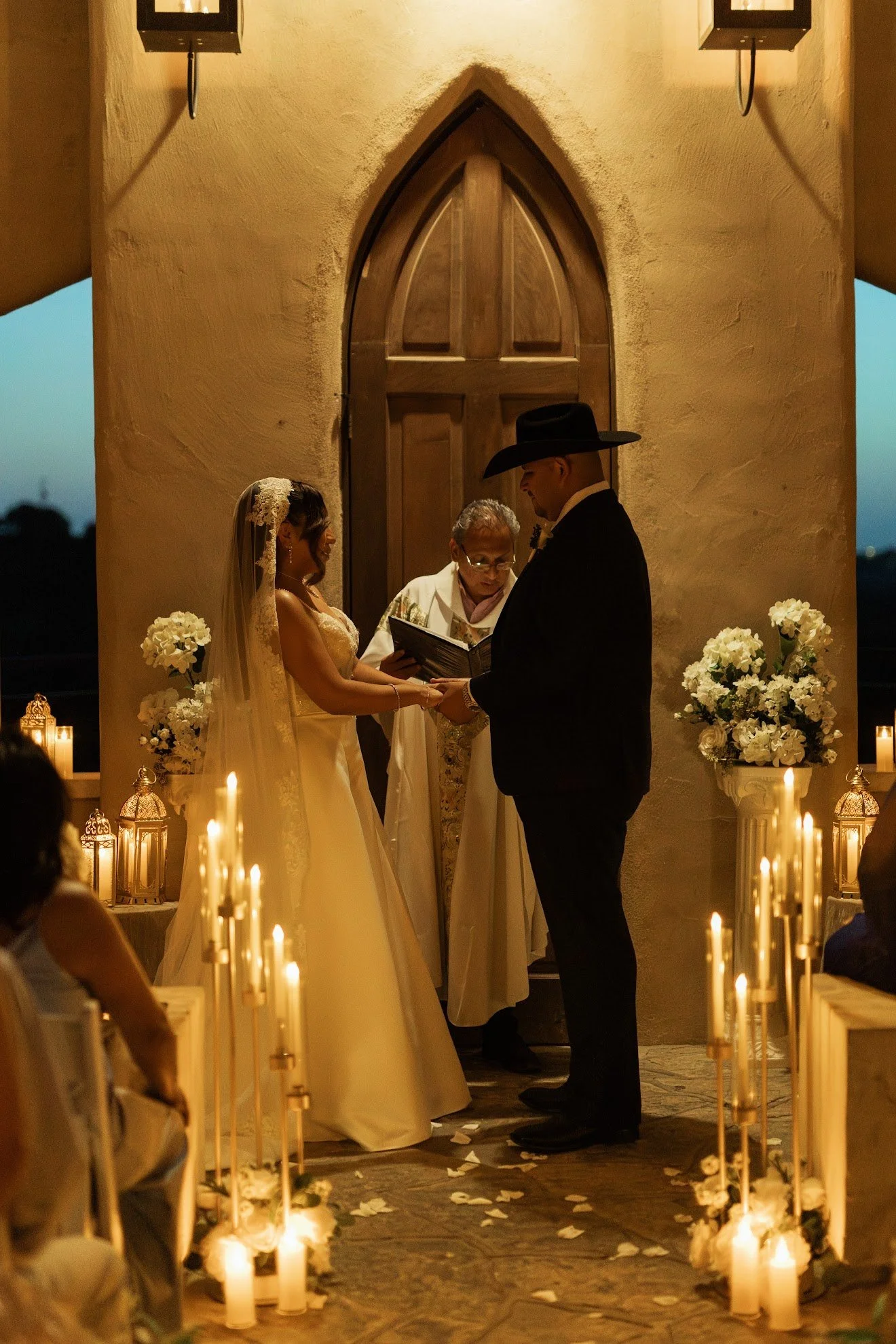 A couple getting married in a church, holding hands and facing each other, with an officiant standing behind them, surrounded by candles and flowers during a candlelit wedding ceremony.