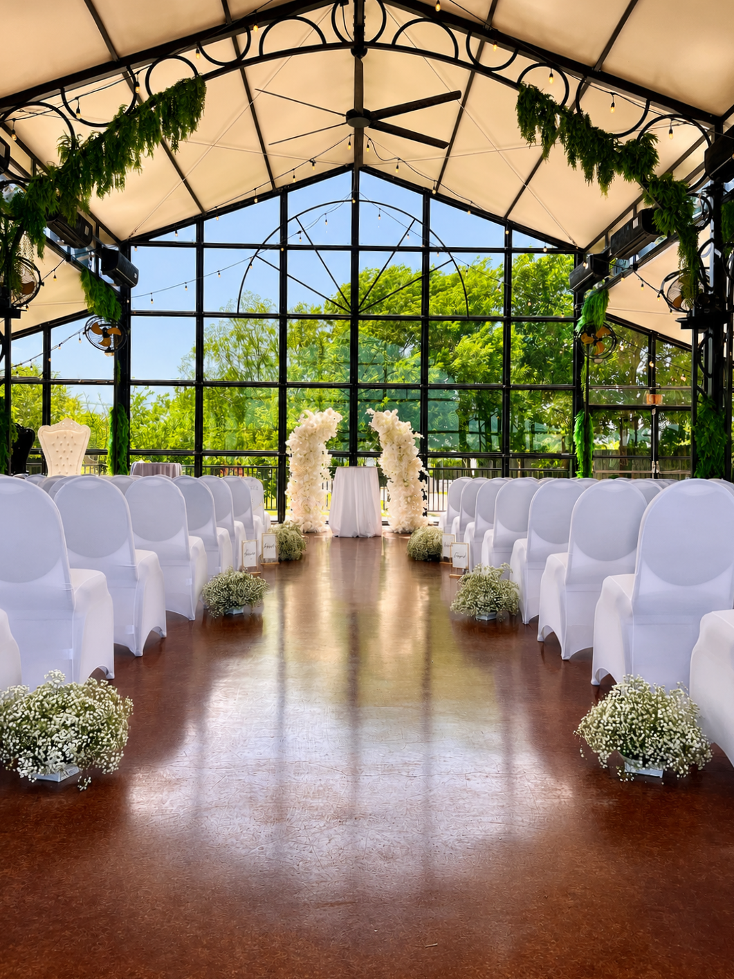 Indoor wedding ceremony setup with white chairs, floral arrangements, and a glass-enclosed backdrop surrounded by greenery.