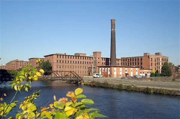Historic brick factory building with a tall smokestack, beside a river with yellow leaves in the foreground under a clear blue sky.