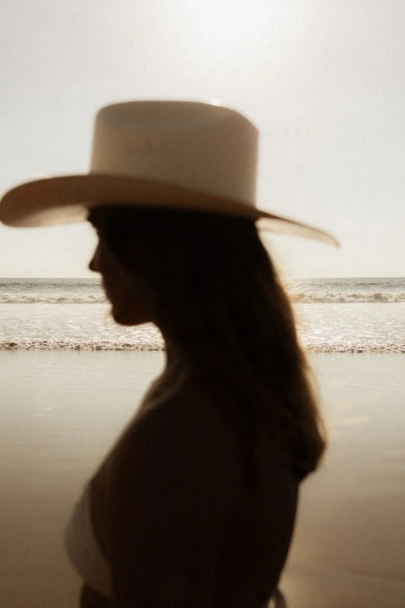 A silhouette of a woman wearing a wide-brimmed hat, standing on a beach with the ocean in the background.