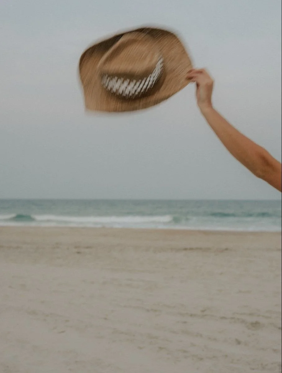 A person holding a straw hat with a smiley face design on a beach with sand and ocean waves in the background.