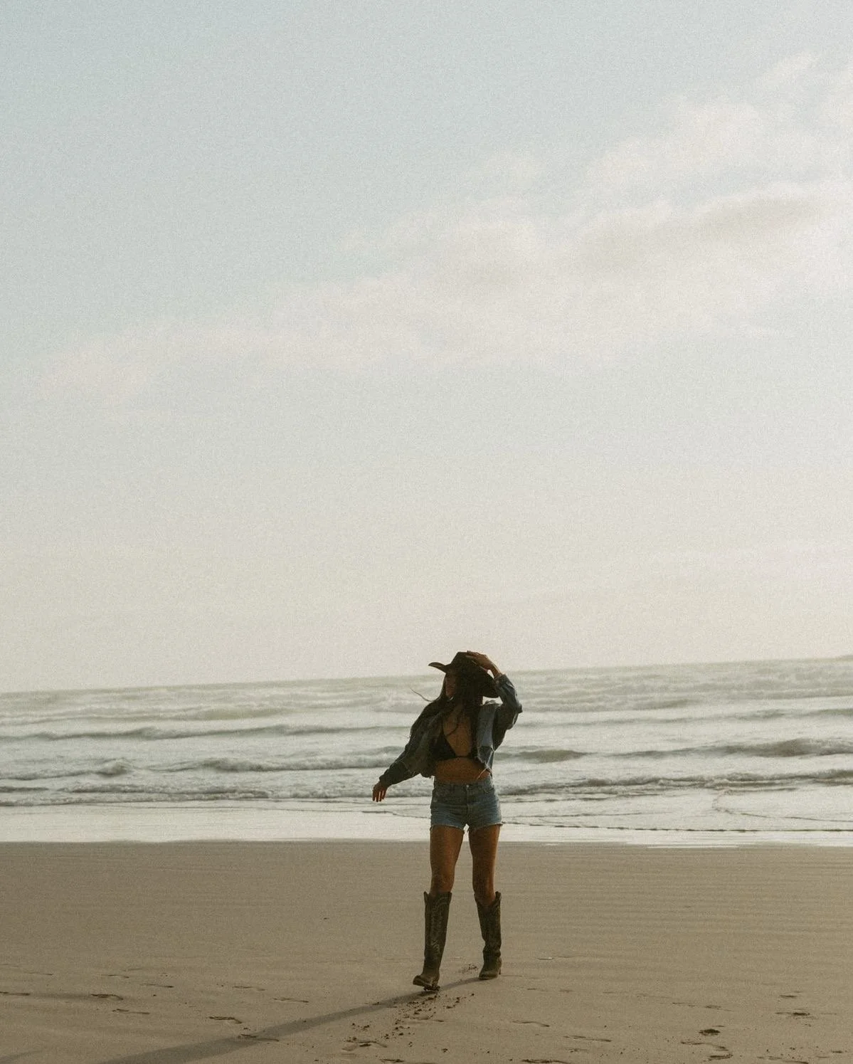 A woman wearing a cowboy hat, denim shorts, a black crop top, a leather jacket, and cowboy boots walking on the beach near the ocean during overcast weather.