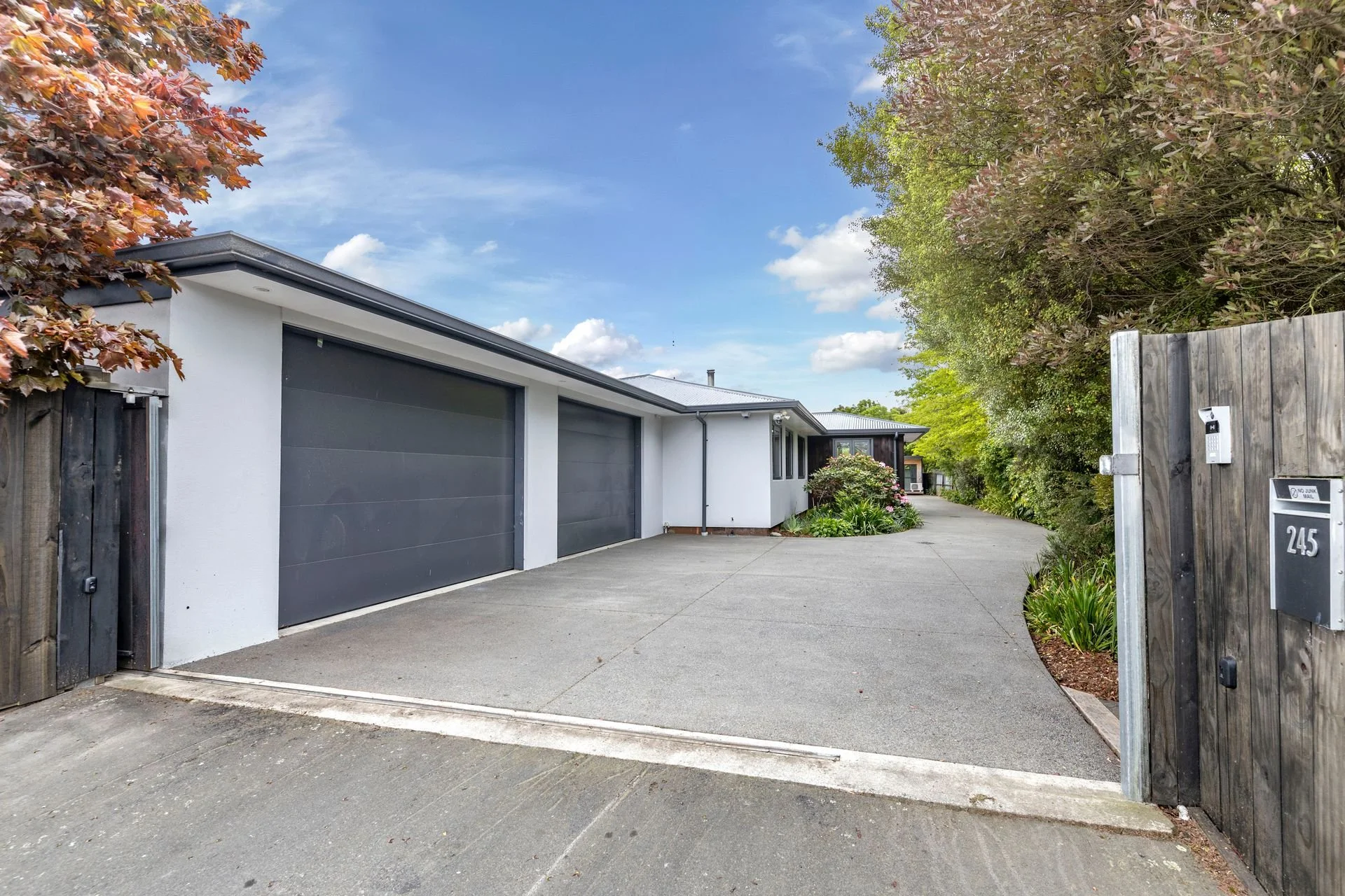 A modern residential house with two large gray garage doors, white exterior walls, and a concrete driveway. There are trees with red and green leaves on either side of the driveway and a wooden fence with a mailbox number 245 on it.