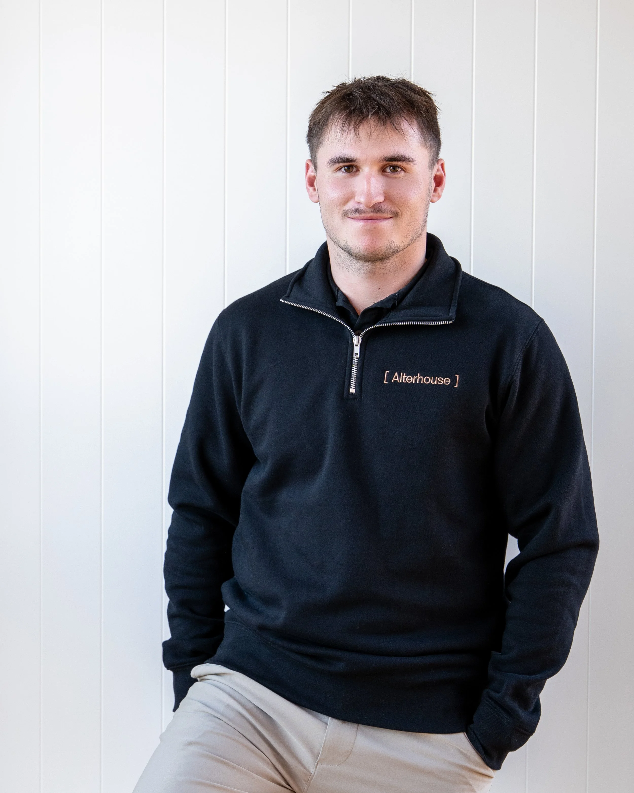 A young man with brown hair and a light beard, wearing a black zip-up jacket with '[Alterhouse]' embroidered on it, standing against a plain white wall. Renovations, Extensions & Additional Dwellings Canterbury | Alterhouse