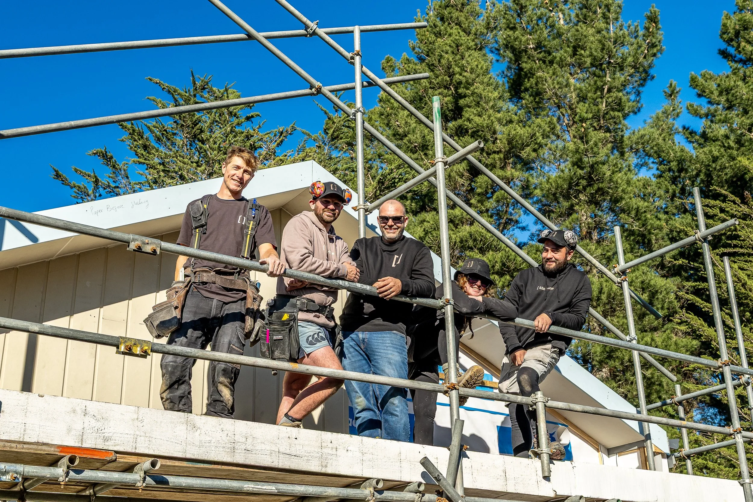 Four people standing on scaffolding in front of a house under construction. They are smiling, wearing work gear, and are outdoors with trees and a blue sky behind them. Renovations, Extensions & Additional Dwellings Canterbury | Alterhouse