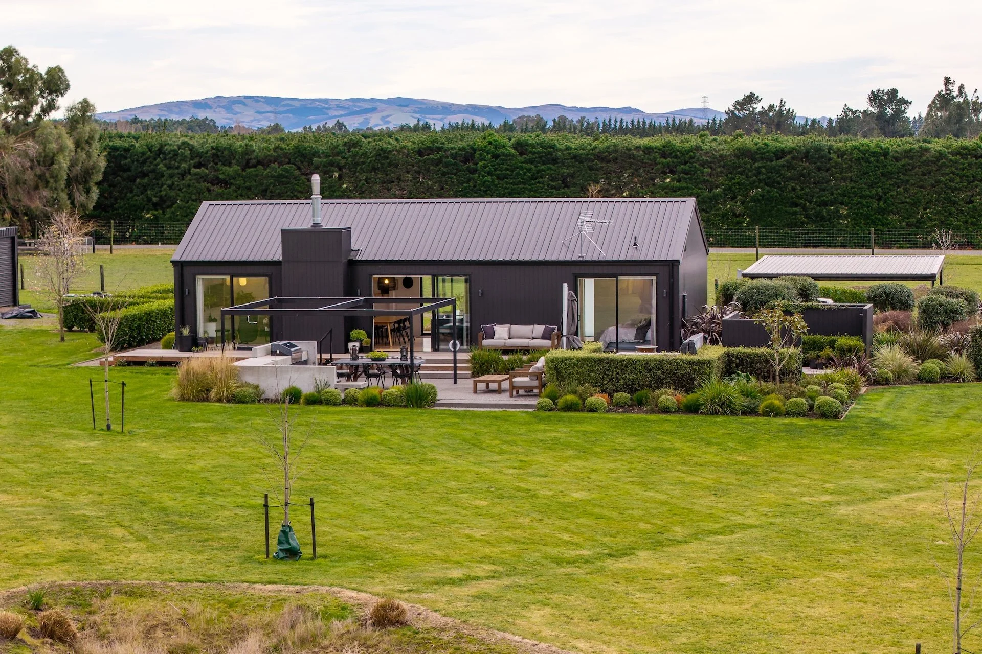 A modern house with large glass doors and a metal roof is set in a lush green landscape with a large lawn, trees, and distant hills in the background. Renovations, Extensions & Additional Dwellings Canterbury | Alterhouse