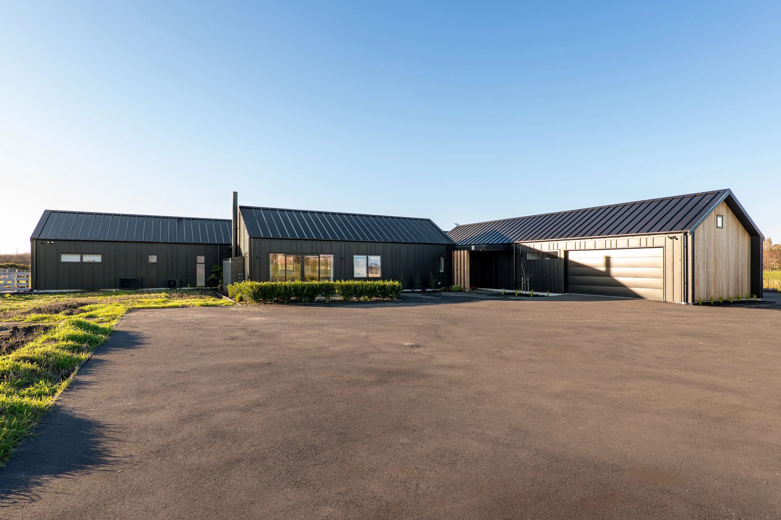 Modern house with black metal roof, wooden siding, large windows, attached garage, and landscaped front yard under a clear blue sky. Renovations, Extensions & Additional Dwellings Canterbury | Alterhouse