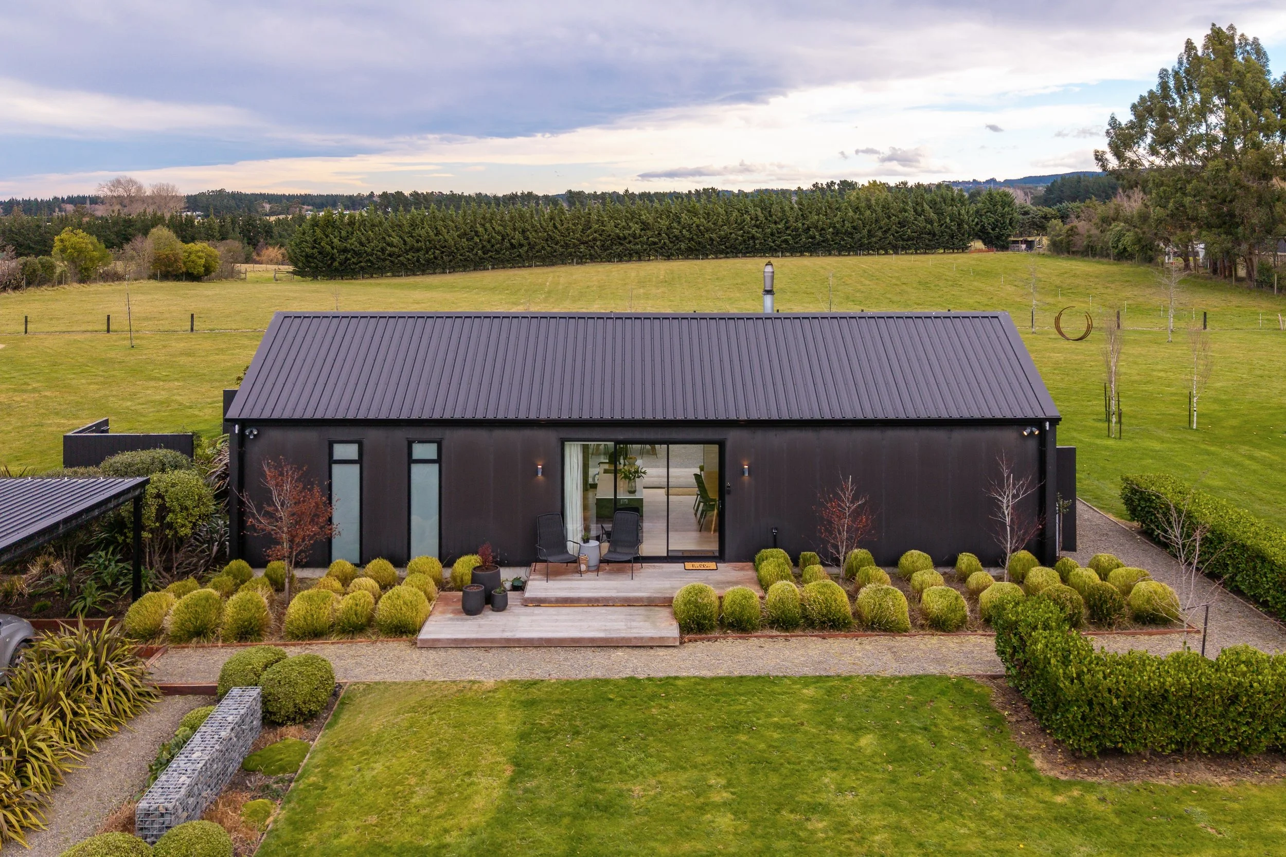 Modern black house with large glass doors, front porch, surrounded by a landscaped garden with trimmed bushes, on a lawn, with trees and fields in the background under a cloudy sky. Renovations, Extensions & Additional Dwellings Canterbury | Alterhou