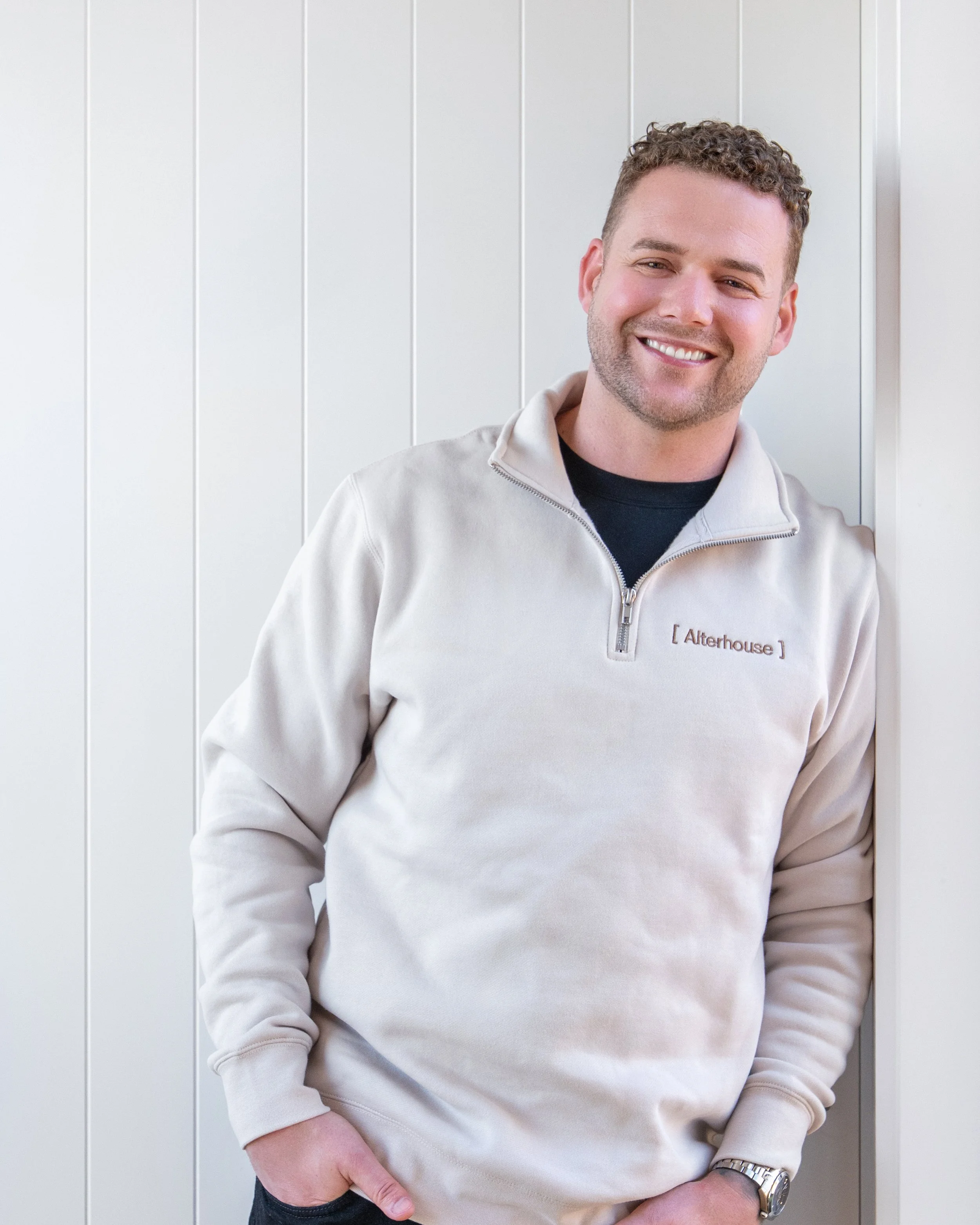A smiling man with short curly hair standing against a white wall with vertical panels. He is wearing a beige quarter-zip sweatshirt with a logo that says 'Alterhouse'. Renovations, Extensions & Additional Dwellings Canterbury | Alterhouse