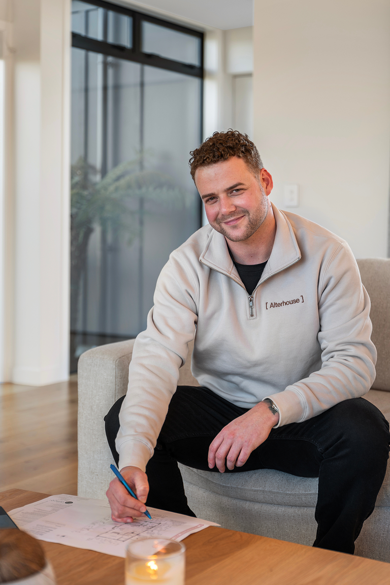 A man with curly hair and a beard sits on a beige couch, smiling, passing a blue pen over some papers on a wooden table. Renovations, Extensions & Additional Dwellings Canterbury | Alterhouse