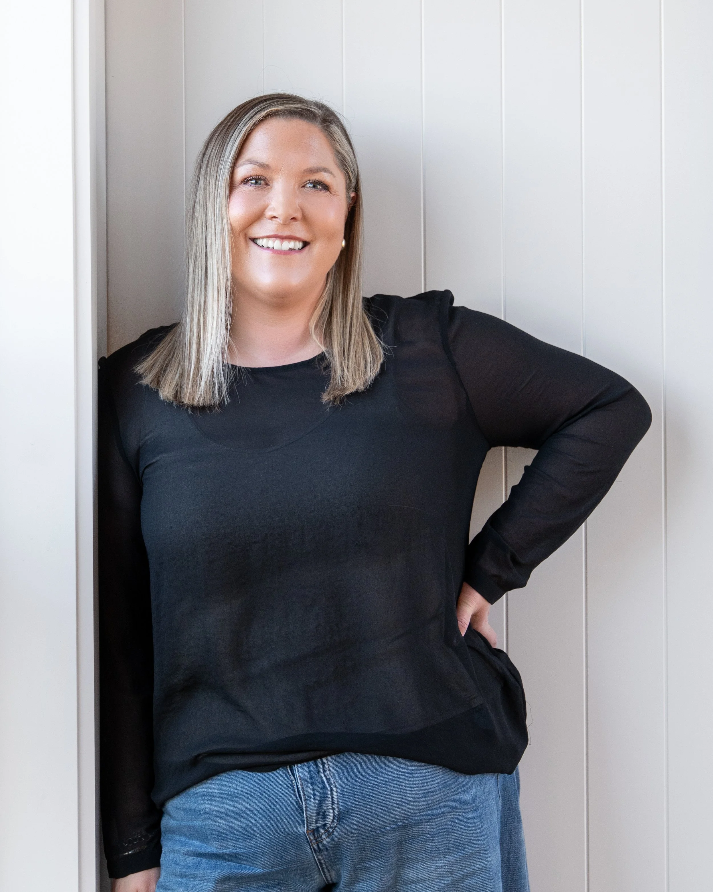 A woman with blonde hair, wearing a black top and blue jeans, standing against a white panel wall, smiling at the camera. Renovations, Extensions & Additional Dwellings Canterbury | Alterhouse