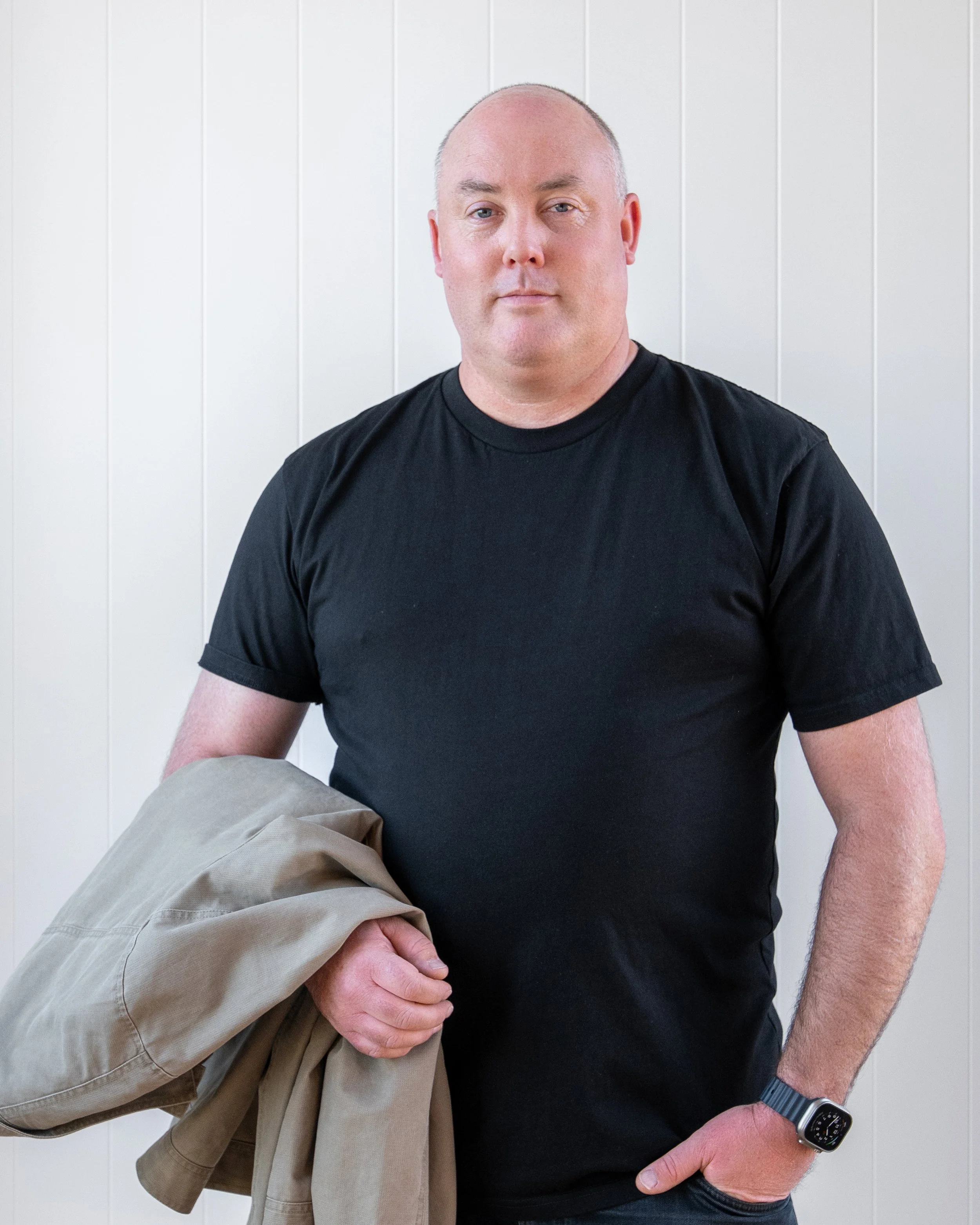 A man wearing a black T-shirt and a wristwatch holds a beige jacket over his shoulder while standing against a white paneled wall. Renovations, Extensions & Additional Dwellings Canterbury | Alterhouse