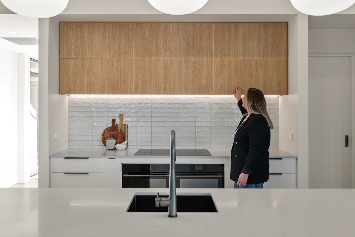 A woman in a black blazer reaches for items on a wooden cabinet in a modern kitchen with white countertops, white subway tile backsplash, and light wood upper cabinets. Renovations, Extensions & Additional Dwellings Canterbury | Alterhouse