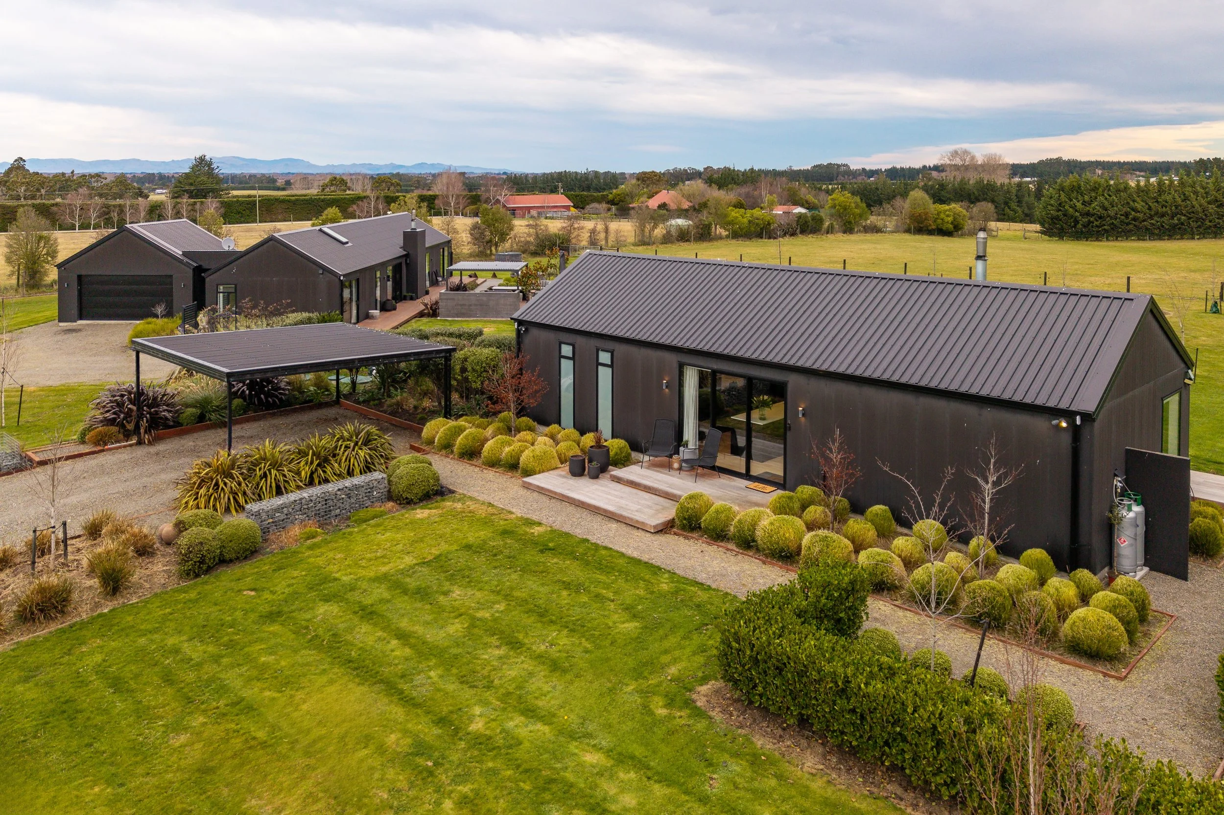 Aerial view of a modern black house with a metal roof, patio, landscaped garden, and green lawn, surrounded by open fields and trees under a cloudy sky.