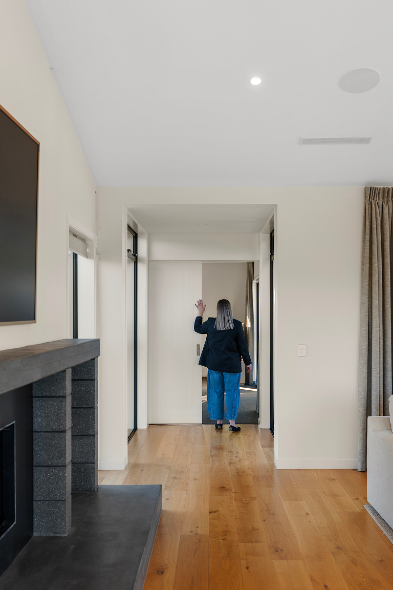 A woman with gray hair wearing a black blazer and blue jeans walking toward a door, with her hand raised in a wave, inside a modern, minimalistic home. Renovations, Extensions & Additional Dwellings Canterbury | Alterhouse