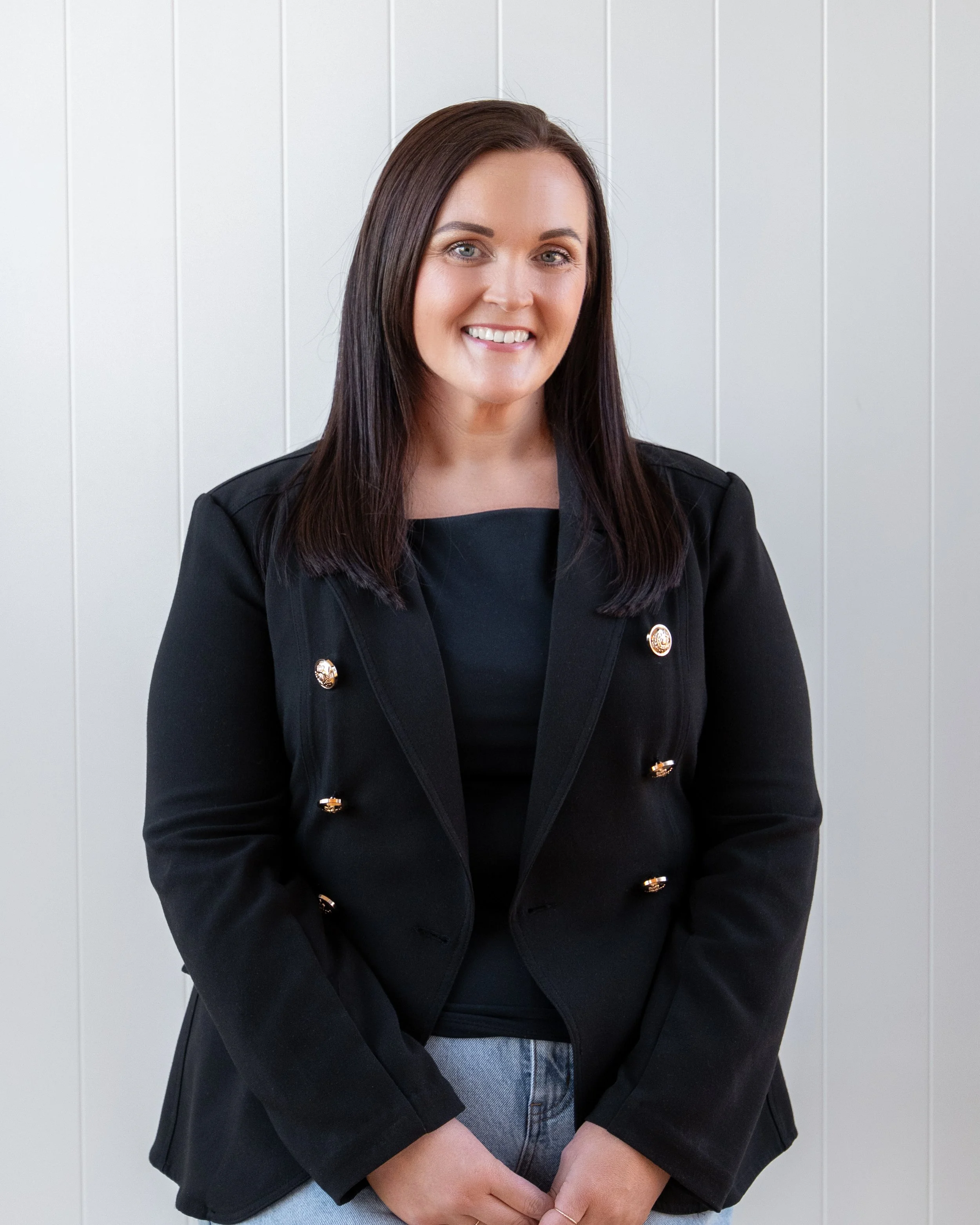 A woman with long dark hair, smiling, wearing a black blazer with gold buttons, a black top, and light blue jeans stands against a white paneled background. Renovations, Extensions & Additional Dwellings Canterbury | Alterhouse