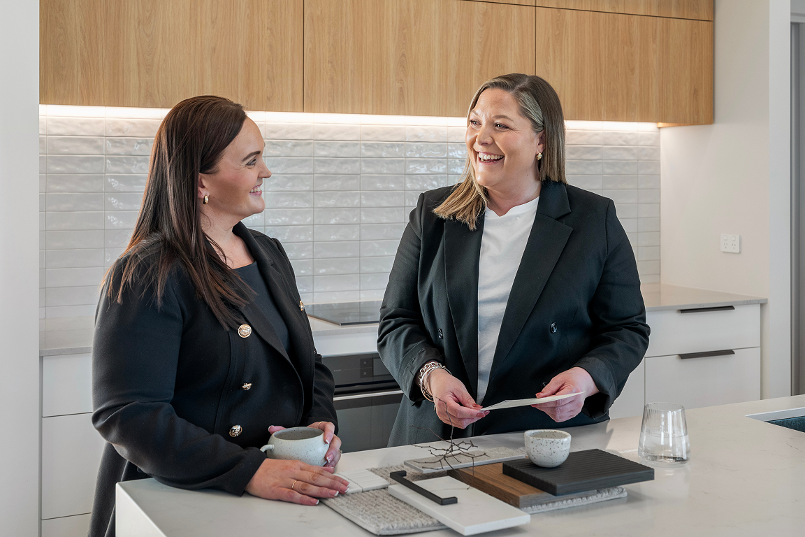 Two women smiling and talking in a modern kitchen, one holding a ceramic cup and the other holding a paper, with various kitchen items on the counter. Renovations, Extensions & Additional Dwellings Canterbury | Alterhouse